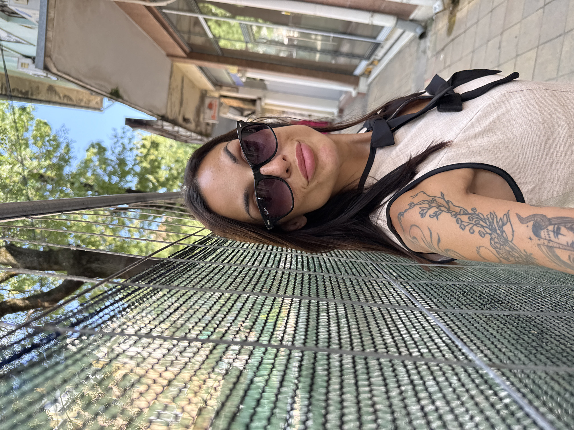 A woman with dark hair, wearing sunglasses and a light-colored dress with black trim and bows, stands in front of a chain-link fence. She has tattoos on her arm. The scene appears to be an outdoor alleyway in the city of Pleven, Bulgaria, with buildings and trees visible in the background. The sky is blue.