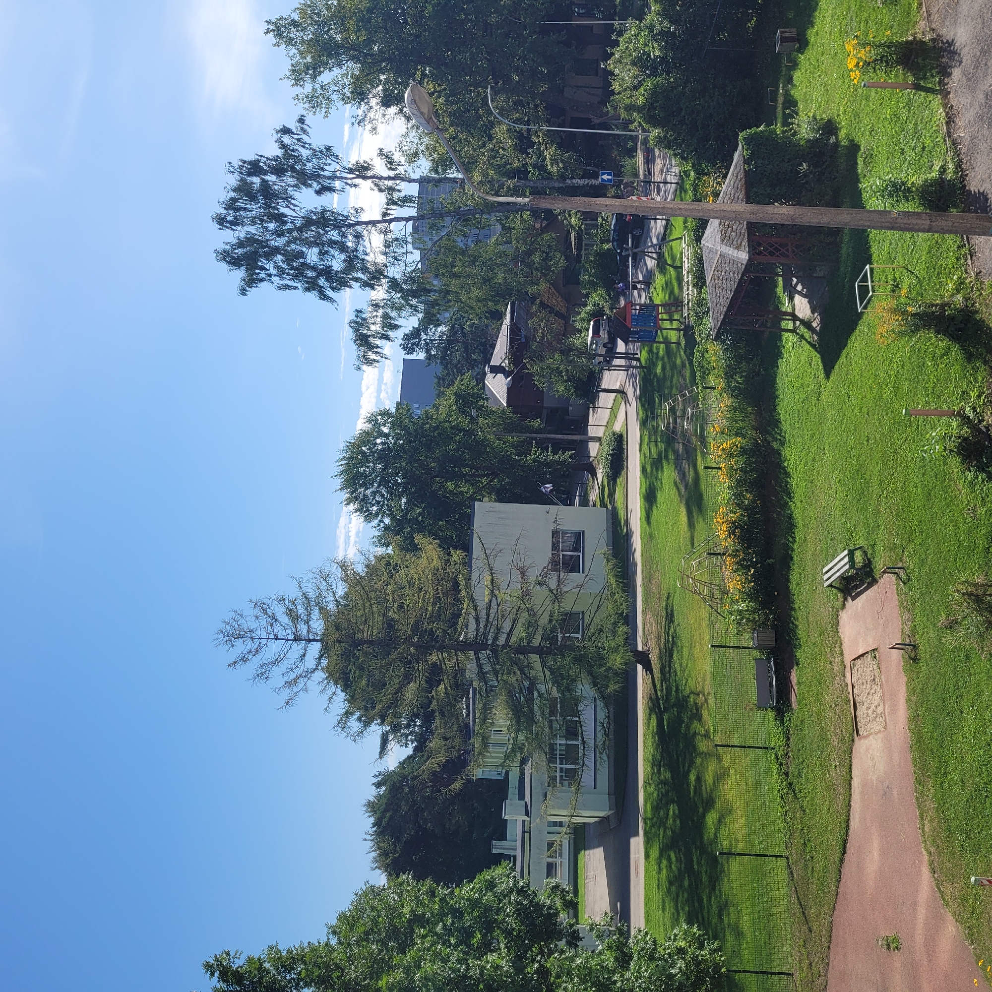 The image depicts an outdoor scene with a building, a playground, and a wooded area. The building appears to be a school or educational facility, with a light green facade and multiple windows. It is situated on a well-maintained lawn. Adjacent to the building is a playground area with climbing structures and swings. A wooden gazebo, partially covered in ivy, stands on the grass. In the background, there are trees and other buildings, suggesting an urban or suburban setting. The sky is clear and blue, indicating good weather. This scene is consistent with a location in Jūrmala, Latvia, known for its schools and recreational areas.