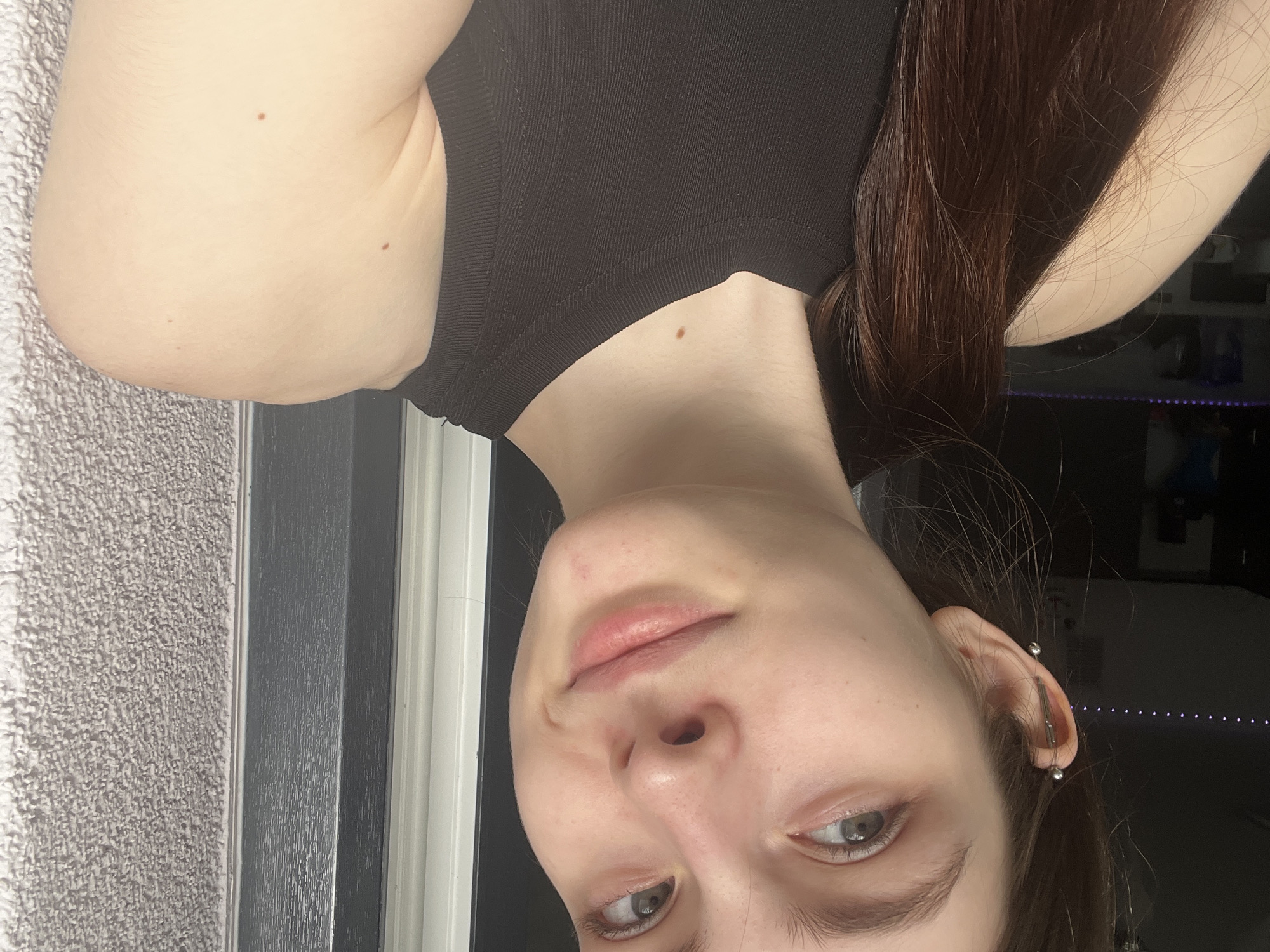 The image is a selfie of a person with fair skin, light eyes, and long brown hair. They are wearing a black tank top. Their ear is pierced with a bar. The background is a mix of a textured wall and a dark room with a glimpse of what appears to be an electronic device with LED lighting. The location cannot be determined from this image alone, therefore the Saint Petersburg location cannot be verified.