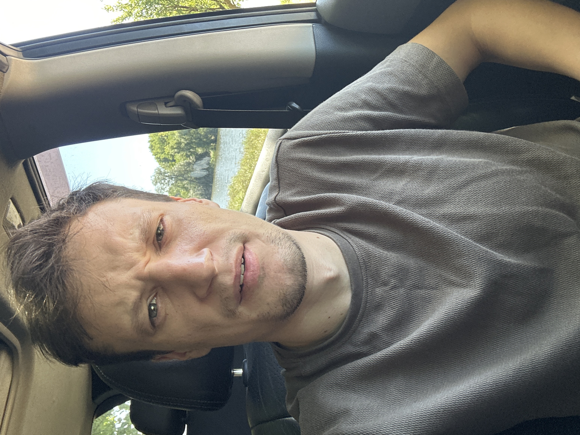 A man is sitting in the driver's seat of a car. He is wearing a grey t-shirt. The car is parked near a body of water, with trees and a blue sky visible in the background. The man is looking at the camera with a slightly furrowed brow. The scene appears to be set in Makiv, Ukraine.