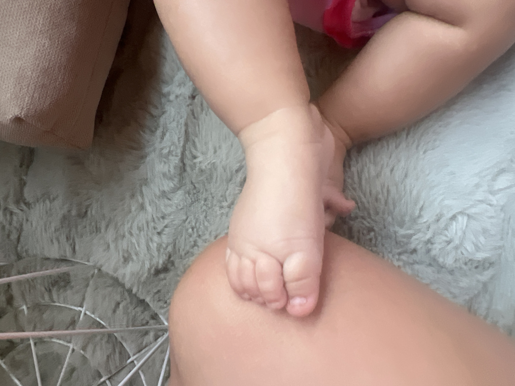 The image shows a close-up of a baby's foot resting on what appears to be an adult's leg or knee. The baby's skin is fair and soft, with clearly defined toes and toenails. The foot is positioned on a textured, fuzzy surface, possibly a blanket or rug, in a soft gray hue. To the left of the baby's foot, a corner of a textured, light brown object is visible, possibly a pillow or cushion. In the upper right quadrant of the image, parts of the baby's legs are visible, with a hint of pink clothing. The overall lighting suggests an indoor setting, likely during the daytime. The scene evokes a sense of tenderness and intimacy, focusing on the delicate details of a baby's foot. There is no visible text in the image. The image was taken in Plovdiv, Bulgaria.
