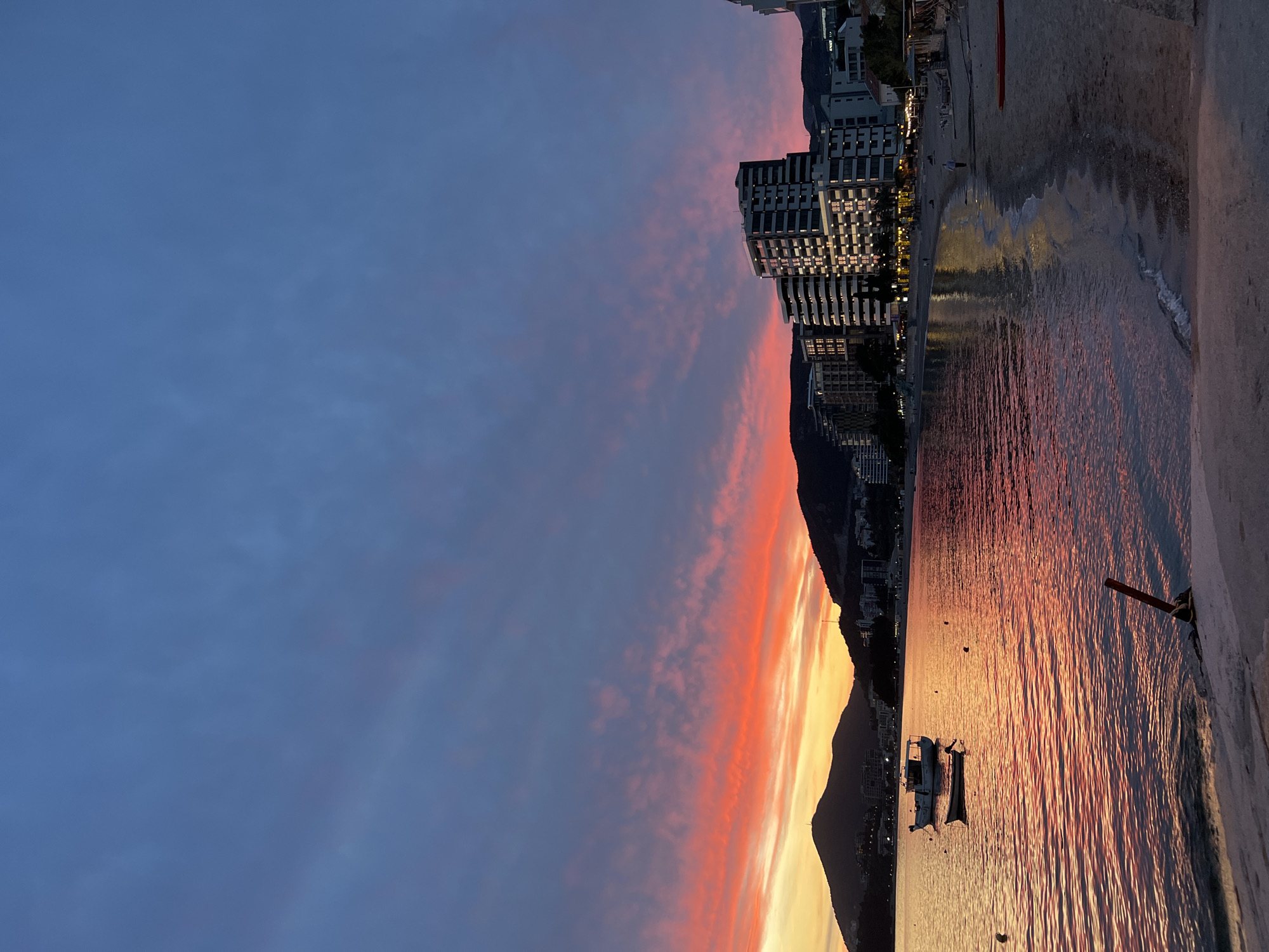 The image captures a scenic view of Rafailovići, Montenegro, during a vibrant sunset. The sky transitions from a deep blue to hues of pink, orange, and yellow, creating a dramatic gradient. Tall buildings line the coastline, reflecting the warm sunlight. A mountain range forms a backdrop to the urban development. The sea mirrors the colorful sky, with two boats visible on the water. The scene is captured from a slightly elevated position, possibly a promenade or pier, marked by a concrete edge on the right side of the image. The overall atmosphere is serene and picturesque.
