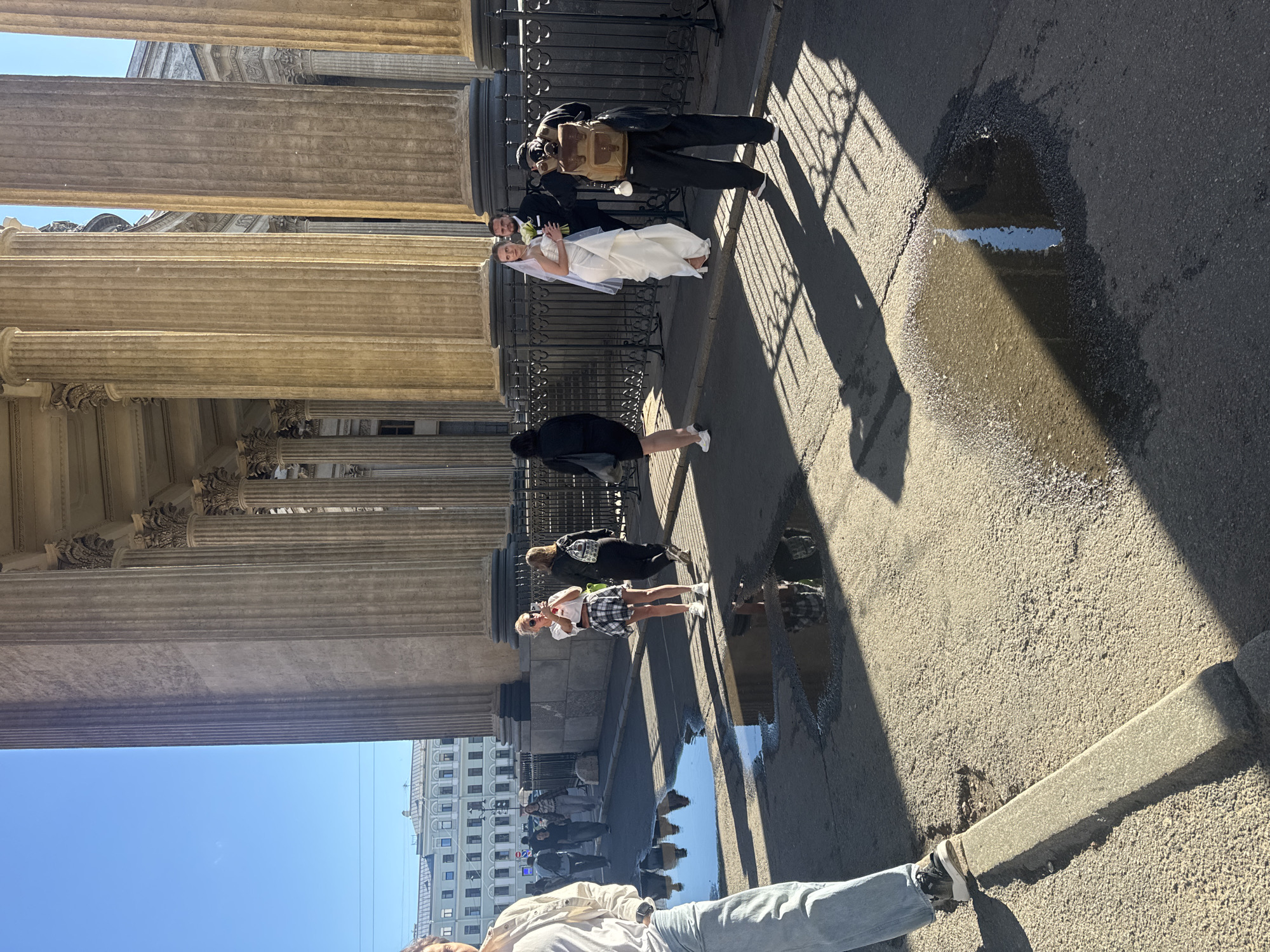 The image depicts a wedding party posing for a photograph in front of the Kazan Cathedral in Saint Petersburg, Russia. A bride in a white wedding dress and veil stands to the right of the groom, who is wearing a dark suit. A photographer with a backpack is positioned in front of them, aiming a camera. Several other people are visible in the background, some walking and others standing, near a fence and large columns of the cathedral. The ground in the foreground is asphalt with several large puddles reflecting the sky and surrounding architecture. The lighting suggests it is daytime with a clear sky, casting strong shadows from the columns and people.