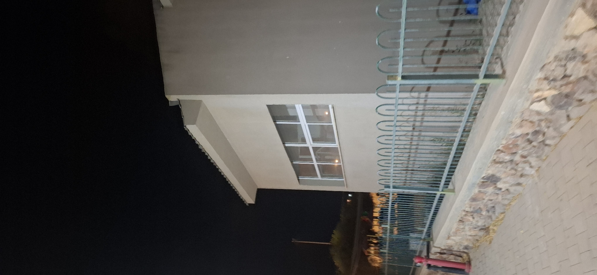 This image captures an exterior view of a building at night, possibly a community center or educational facility, located in Yerocham, Israel. The building features a light-colored facade and a distinctive roofline with visible tiling. A large, horizontal window with multiple panes is a prominent feature of the building's side wall, reflecting some internal lighting. In front of the building, a dark green metal fence with a decorative, arched top runs horizontally, separating the property from a paved walkway. The walkway is constructed of light-colored rectangular pavers and bordered by a low wall made of rough-hewn stones. A red fire hydrant is partially visible on the left side of the image, near the walkway. The background is dark, indicating nighttime, with some faint lights and greenery visible in the distance, suggesting a landscape beyond the fence.