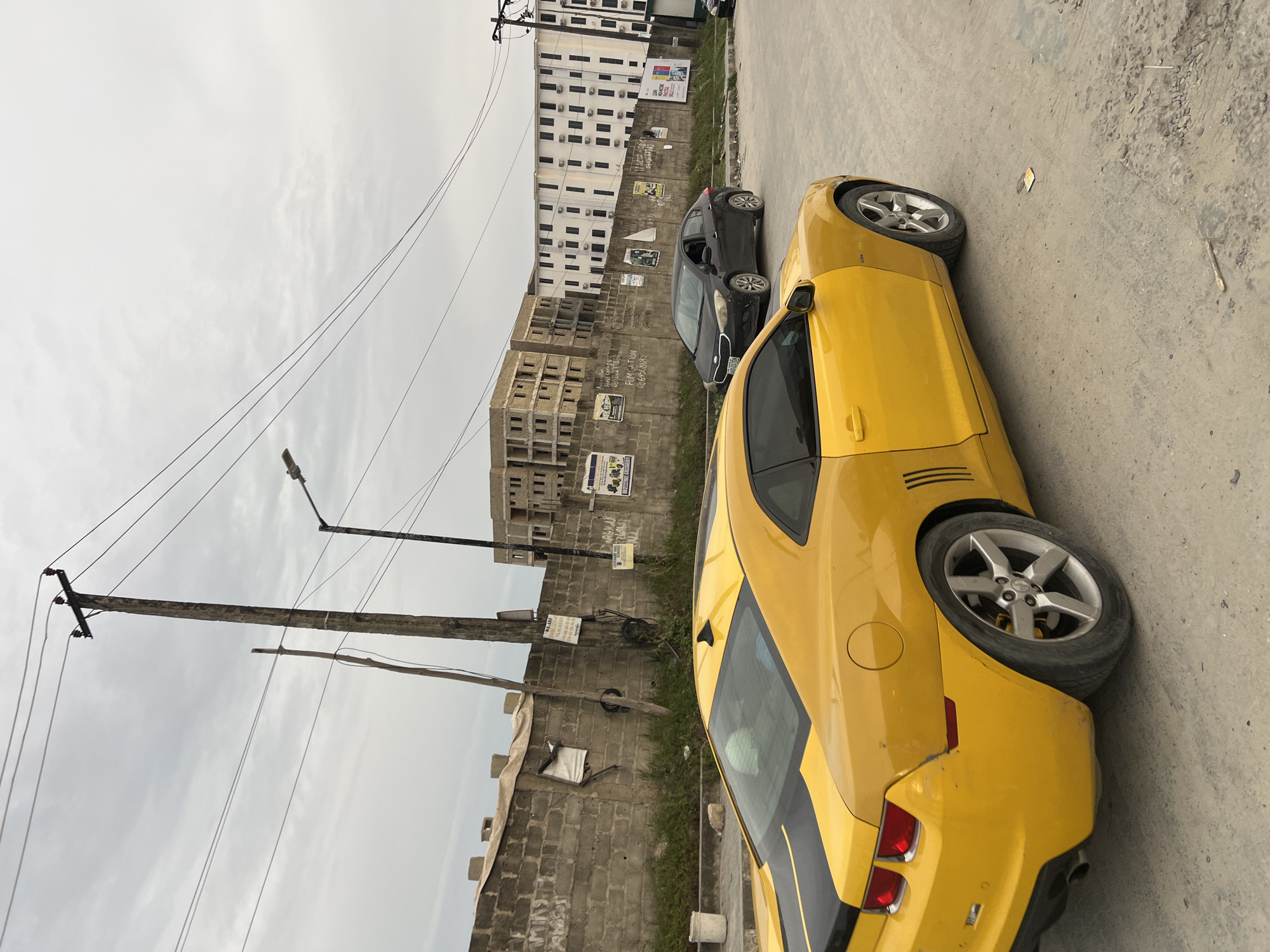 This image presents a street scene in Igboefon, Nigeria, under an overcast sky, indicating daytime with diffused light. The primary subject is a vibrant yellow Chevrolet Camaro sports car, featuring black racing stripes, parked on the right side of a dusty, unpaved road. Its rear section, including the trunk, taillights, and part of the side with a silver alloy wheel, is prominently visible.
Further down the road, a black Kia Rio sedan is also parked. The background reveals a mix of urban structures. On the left, a large, multi-story white building with many air conditioning units appears to be completed, while other buildings constructed from bare concrete blocks seem to be under construction or partially developed.
A low concrete block wall lines the street, serving as a canvas for various advertisements and signs. Visible text includes "FUMIGATION" accompanied by a phone number starting "0806-6268...", and another sign that reads "WE ARE..." with a phone number "080 34 39..." and likely services like "SALES," "RENT," or "LEASE." A larger, partially obscured sign on the white building appears to mention "HEALTHCARE."
Utility infrastructure is a significant element on the left, featuring multiple sturdy wooden poles supporting numerous overhead electrical wires, and a street lamp extending into the sky. No people are visible, and no specific event or activity is taking place; the scene captures a static moment in a developing urban landscape.