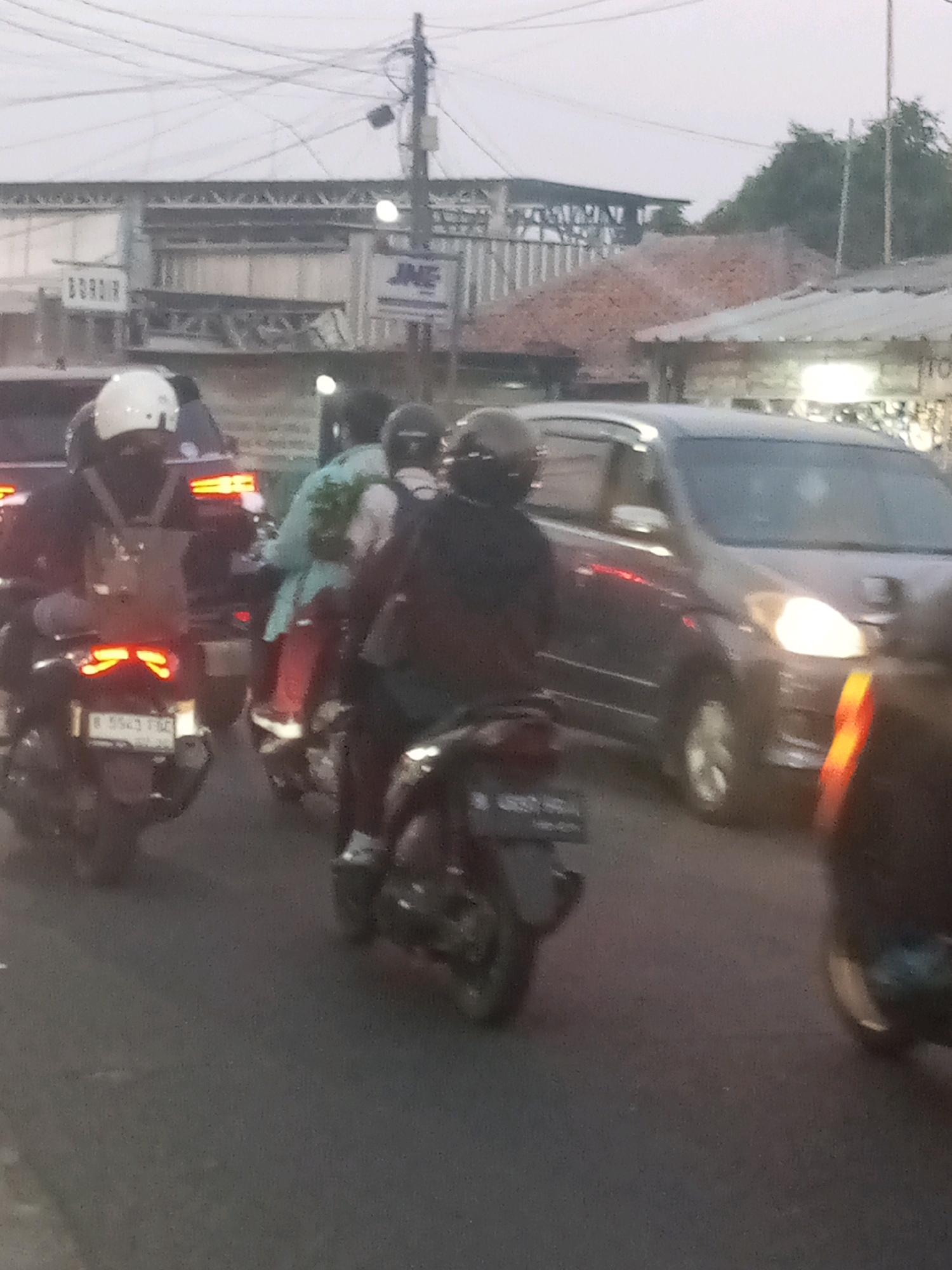 The image depicts a street scene in Bekasi, Indonesia, with several motorcycles and a car on a road, likely during dusk. In the foreground, a person on a motorcycle is visible from behind, wearing a dark jacket and a helmet. To the left, another motorcycle with a white helmet is also seen from behind. Further back, a motorcycle with two occupants, one wearing a light blue shirt and red pants, is present. A dark-colored car is in the background, with its headlights on. Various buildings and structures line the road, including what appears to be a commercial building with signage. The sky is overcast, suggesting a cloudy or hazy atmosphere typical of urban environments. The overall impression is of moderate traffic congestion on a street in an Indonesian city.