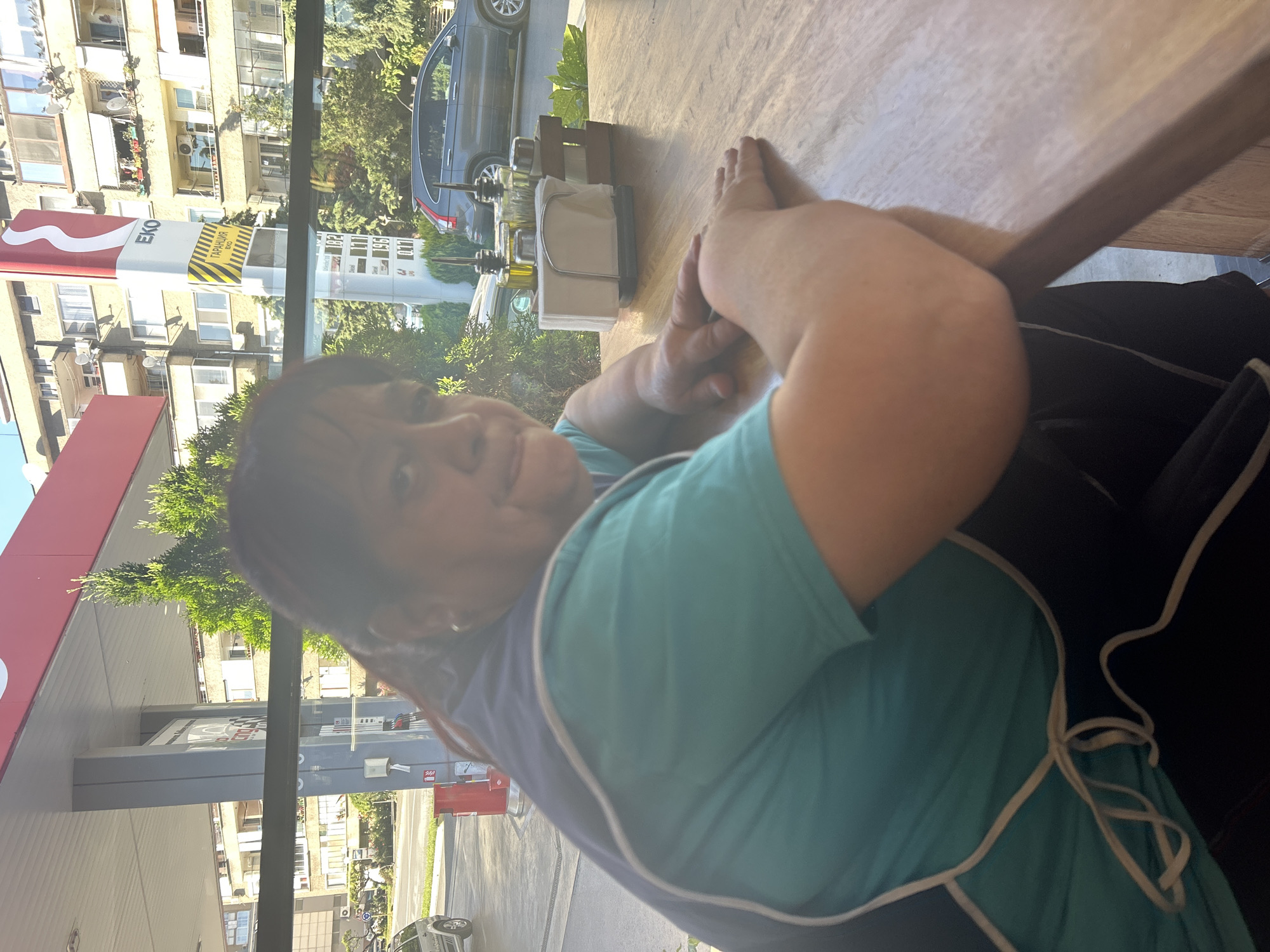 A woman is seated at a table, her hands resting on its surface. She wears a teal shirt with a navy blue apron tied at the waist. Outside the window behind her is an EKO gas station, a parking lot with cars, and several multi-story residential buildings. Based on the architecture and the presence of the EKO gas station brand, the location is likely in Kazanlak, Bulgaria.