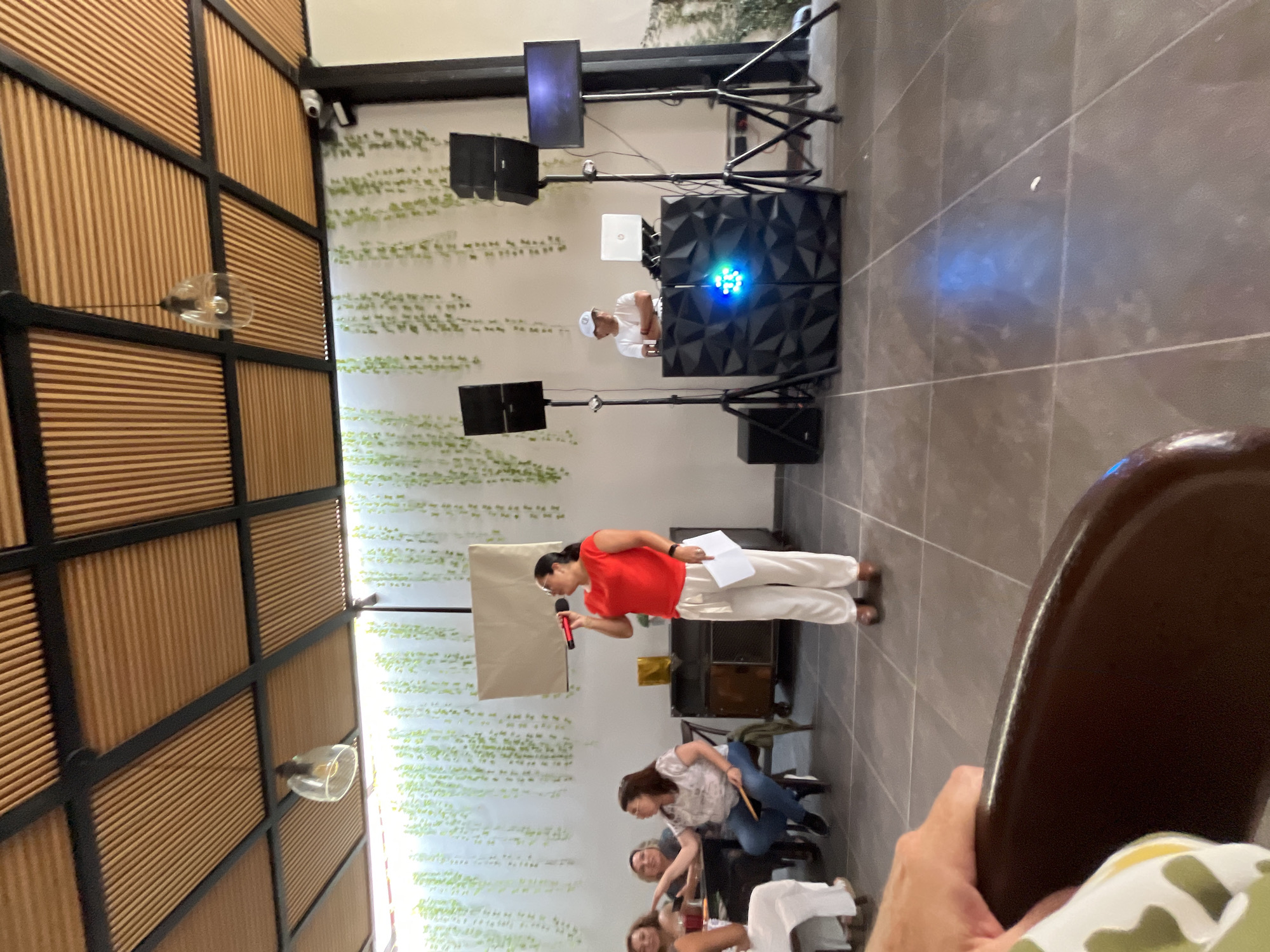 Here's a description of the image:
**Location:** San Bernardino Tlaxcalancingo, Mexico.
**Scene:** A presentation or talk is taking place indoors.
**People:** A woman in a red shirt and white pants is speaking into a red microphone, holding papers in her left hand. She is positioned near a blank presentation board. In the foreground, several people are seated and appearing to listen. Behind the presenter, a person is at a DJ setup, possibly operating sound and lighting equipment.
**Objects:** Presentation board, red microphone, papers, speakers, sound and lighting equipment, tables, chairs.
**Actions/Interactions:** The woman is giving a presentation. The audience is listening. The person at the DJ setup may be managing the sound and visual aspects of the presentation.
**Environment:** The room has a decorative wall with light green plant patterns. The other wall has a grid pattern with wooden planks. The floor is tiled.