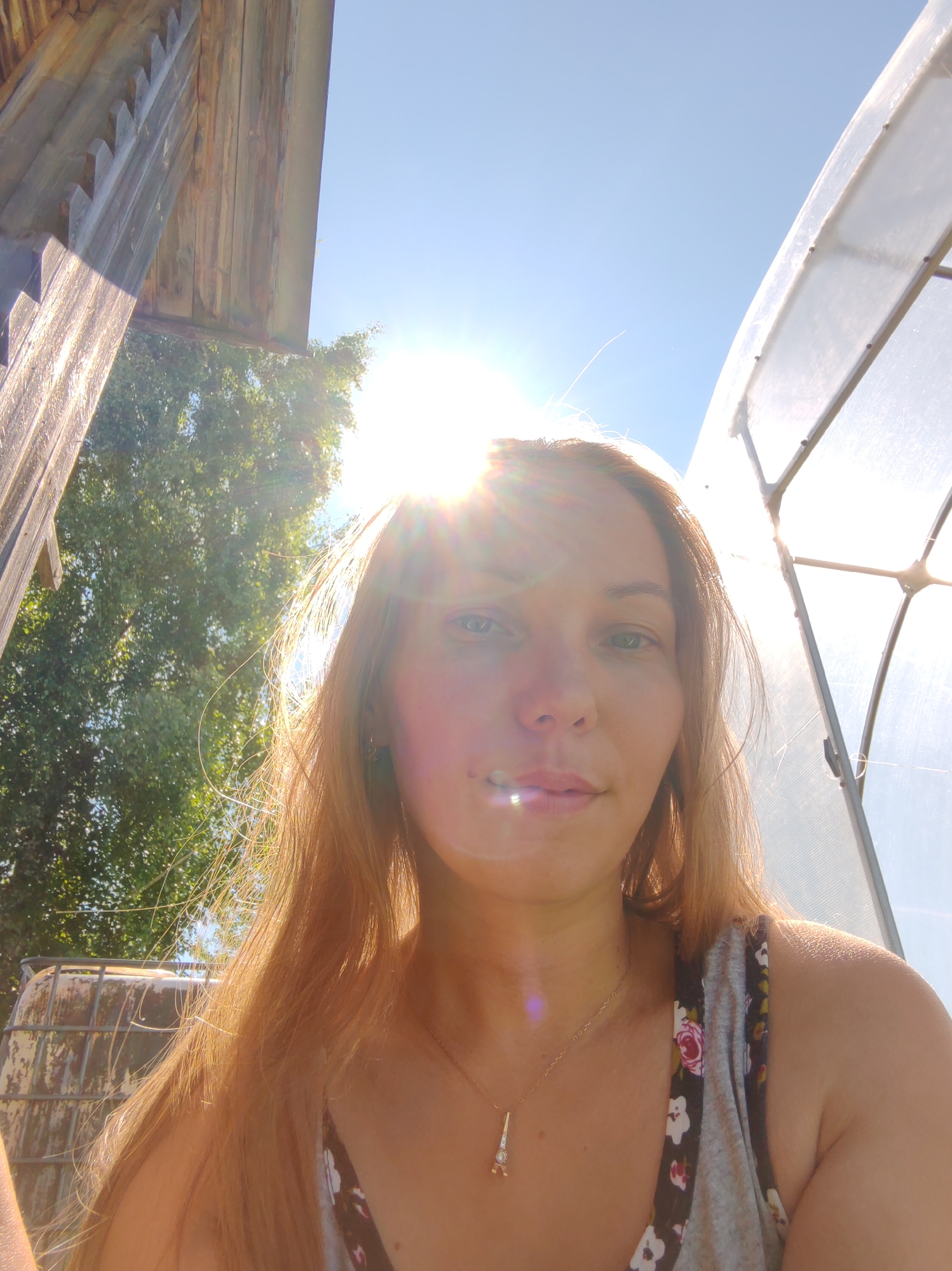 A woman with shoulder-length blonde hair takes a selfie outdoors in what appears to be a rural setting. She is facing forward, and the sun is directly in front of her, creating a lens flare and obscuring parts of her face. She is wearing a grey tank top with a floral pattern. A delicate gold necklace with a charm resembling the Eiffel Tower is visible around her neck.
The background shows elements of a rustic environment. To the left, a portion of a weathered wooden structure with a peaked roof is visible, suggesting it might be part of a barn or outbuilding. Further back, green trees with dense foliage are present, indicating a sunny day with clear skies. To the right of the woman, a large, translucent structure, likely a greenhouse, is partially visible, its curved frame and plastic sheeting illuminated by the sun. In the distance, behind the trees on the left, a rusty metal tank or container can be seen.
The bright sunlight and clear sky suggest it is daytime, likely mid-morning or afternoon, on a clear and warm day. The setting appears to be a private yard or rural property in Чухлэм, Russia, judging by the combination of agricultural structures, vegetation, and the overall atmosphere. The woman's expression is neutral, though the strong sunlight makes it difficult to discern fine details. There is no visible text in the image.