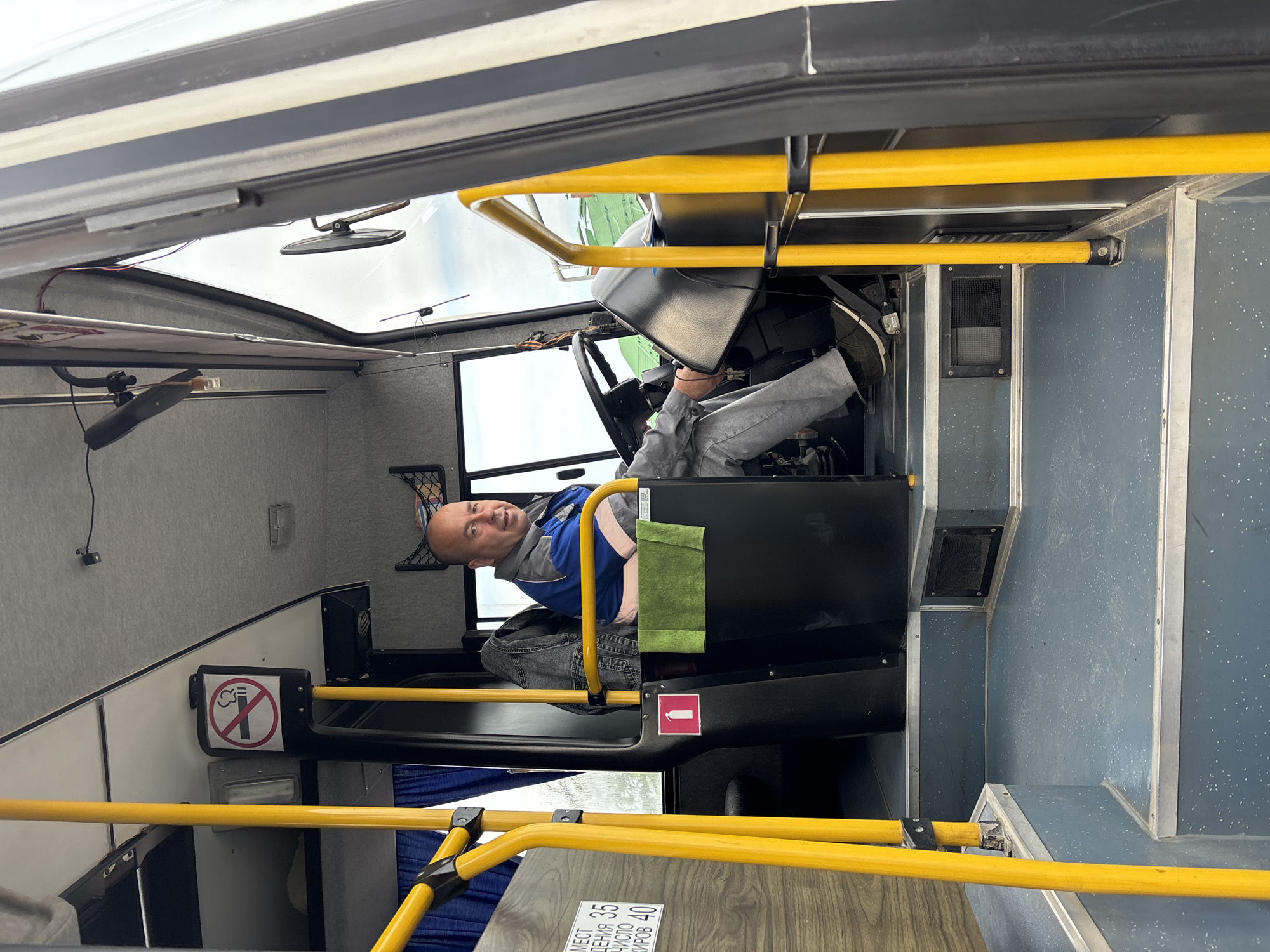 The image displays the interior of a public transport vehicle, likely a bus, with the driver in the foreground. The photo is oriented ninety degrees clockwise.
**Subjects:** The main subject is a man, presumably the bus driver, seated in the driver's cabin. He is a middle-aged man with short hair, looking directly at the camera with a slight, friendly smile. He wears a blue and grey uniform jacket over a lighter shirt and grey trousers. The vehicle's interior features numerous yellow grab rails, a black driver's console with a steering wheel, and a functional-looking cabin. The walls are light grey with a textured pattern, while the floor and lower panels are blue.
**Activity/Event:** The driver is seated at the controls of the bus, appearing to be stationary, possibly waiting or on a break, as he is making eye contact with the person taking the photo.
**Notable details:** Through the front windshield, an overcast sky is visible, suggesting daytime. A small rearview mirror is attached near the top of the windshield. A green cloth or towel hangs over a part of the driver's console. Blue curtains are drawn on one side window. The overall setting is utilitarian, typical of public transportation. Given the provided context, the location is Chaikovsky, Russia.
**Visible text:** A prominent "No Smoking" sign, depicted by a crossed-out cigarette icon within a red circle, is visible near the driver's seat. Below it, a red square with a white fire extinguisher icon indicates the location of safety equipment. On a wooden-patterned surface near the front (likely a floor panel or part of the dashboard), partially visible Russian text reads "МЕСТ," "ДЕНИЯ 3.5," "СЛО 40," and "ИРОВ 40," which likely pertains to the seating and total capacity of the vehicle, for example, "Мест для сидения 35, Общая вместимость 40 пассажиров" (Seating capacity 35, Total capacity 40 passengers).