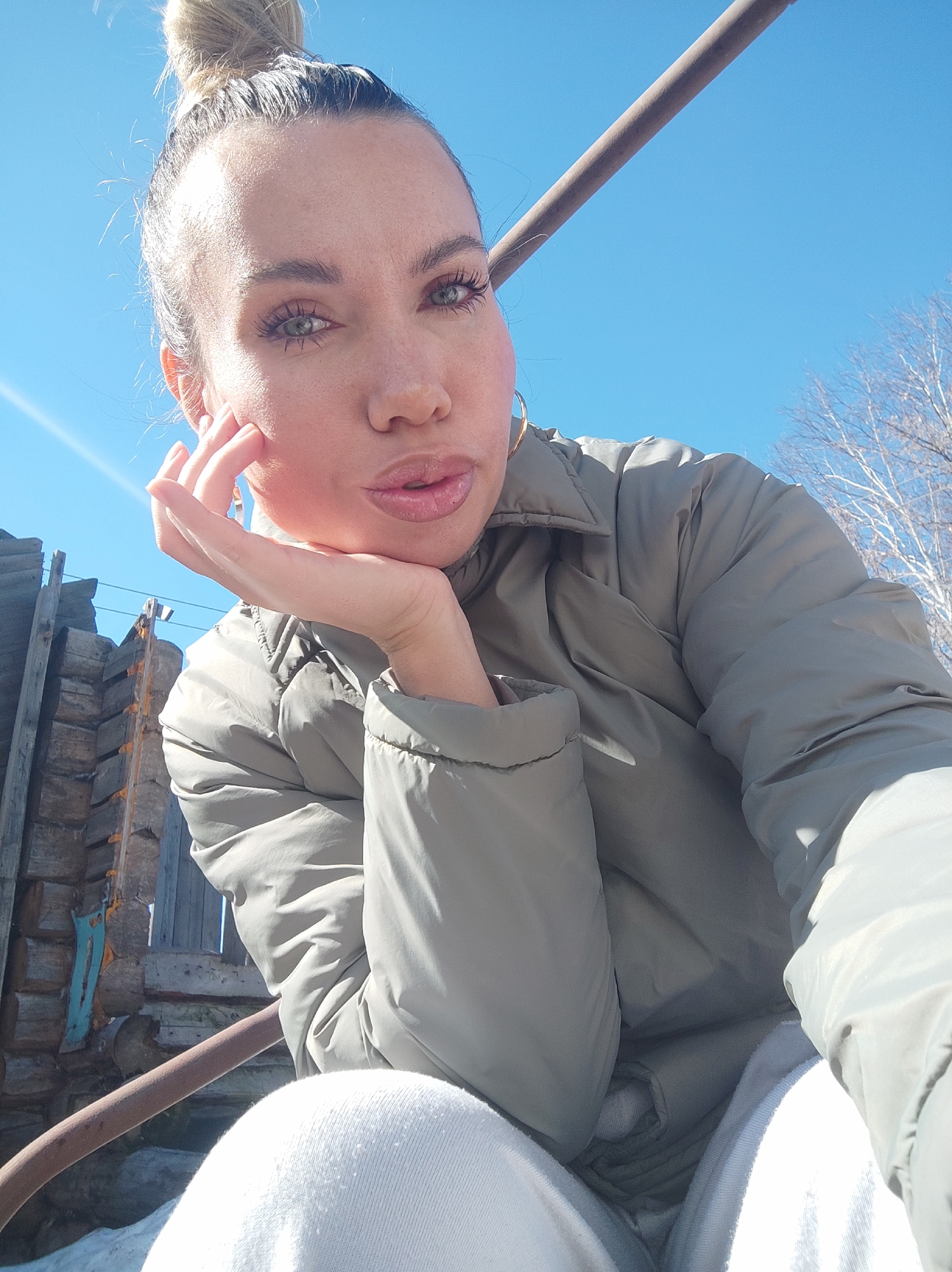 The image is a selfie of a woman with blonde hair in a bun. She is wearing a light gray puffer jacket and white pants. She is holding her face with her hand and looking at the camera. The background shows a clear blue sky, bare trees, a wooden structure, and what appears to be a snowy ground. Without specific geolocation data, it is not possible to confirm or deny that it was taken in Votkinsk, Russia.