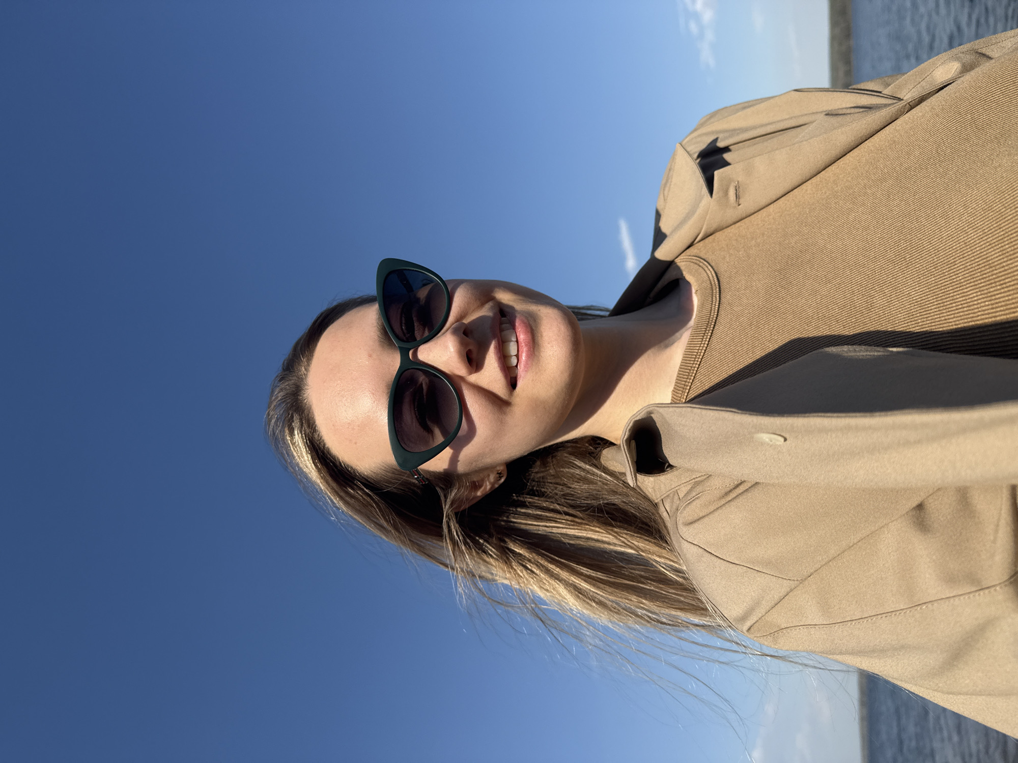 A woman stands outdoors against a bright blue sky, wearing sunglasses with dark lenses and green frames. She has fair skin and blonde hair, which is blowing slightly in the wind. She is wearing a tan-colored top and a trench coat of the same color. The location appears to be in the city of Chaikovsky, Russia, as indicated by the presence of a body of water in the background, possibly the Kama Reservoir.