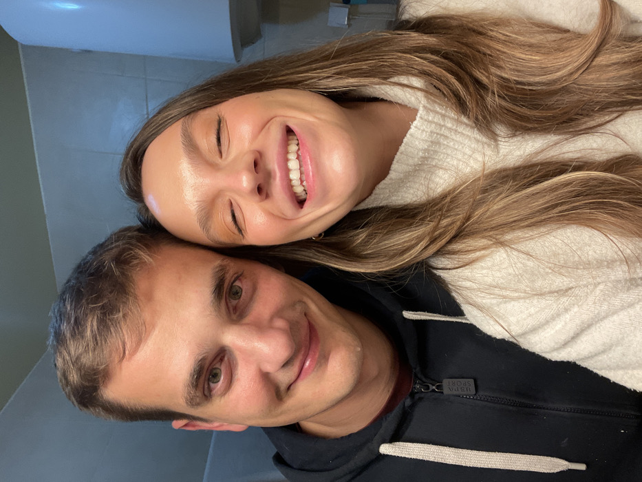 A rotated selfie features a smiling couple. The woman above has light brown hair and a bright smile, wearing a white sweater. Below, the man smiles with his head close to hers, wearing a dark zip-up hoodie. The background includes a wall, possibly tiled. The image appears to be taken indoors with soft lighting.