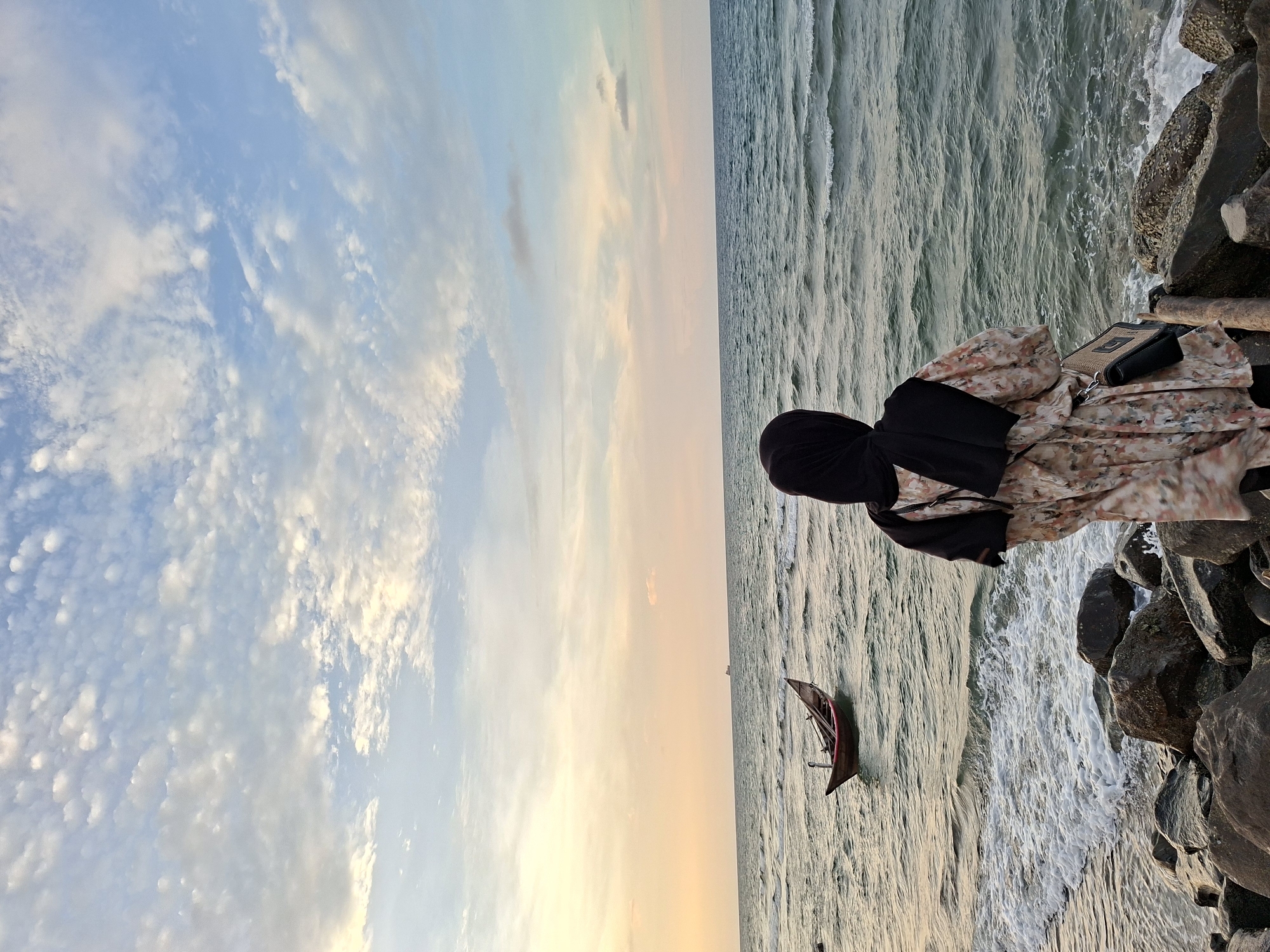 A person wearing a hijab and floral patterned clothing is standing on rocks at a beach. The sea is to the right and the sky is to the left. A small boat is in the water. The sky is cloudy with colors of blue, white, and orange from the sunset. The location is likely in Lhokseumawe, Indonesia.