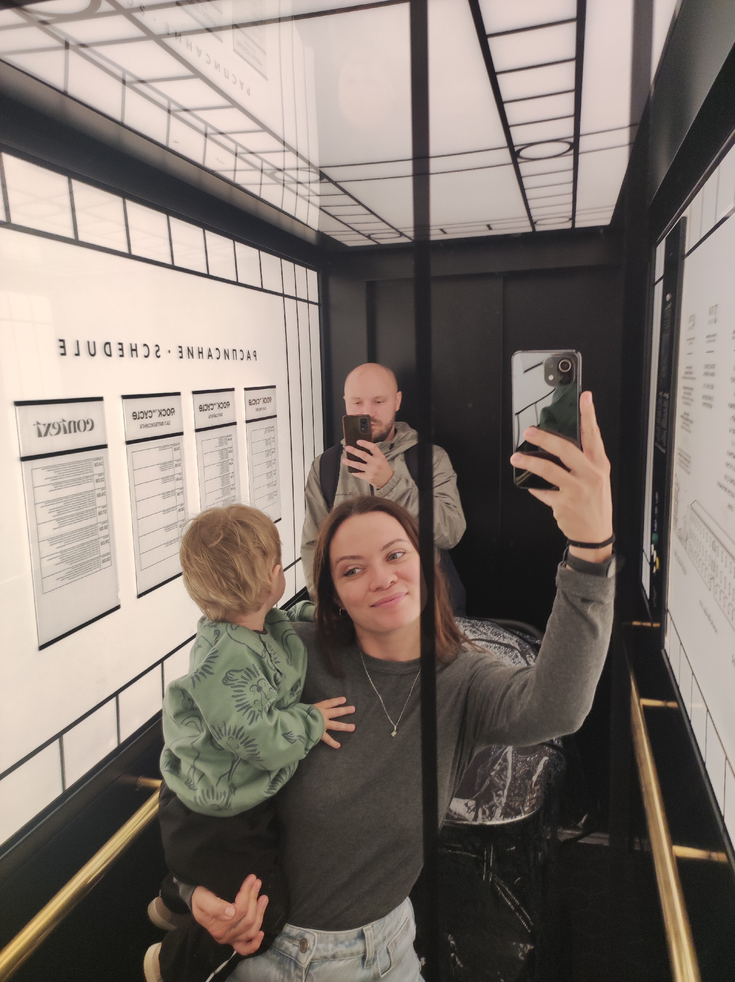 The image shows a family of three taking a selfie in a modern elevator, likely in Saint Petersburg, Russia, given the Russian text visible on the wall. In the foreground, a woman is holding a young child on her left arm while holding her phone in her right hand to capture the reflection. The child is wearing a green hoodie with a pattern. Behind them, reflected in the elevator mirrors, a man is visible, also taking a picture with his phone. The elevator walls are black with white panels displaying text, including what appears to be a schedule. The ceiling is illuminated with a grid of lights. A gold handrail runs along the bottom of the elevator.
