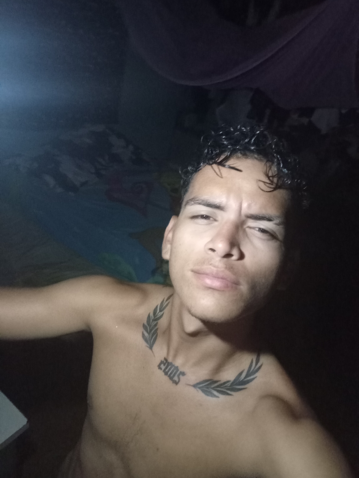 A dimly lit, close-up photo captures a shirtless, young man with curly black hair and a tattoo of laurel branches with the number "2005" on his chest. He appears to be indoors, with the background showing what looks like a bed covered with sheets and draped fabric. The specific location within Altagracia, Venezuela, cannot be determined from the visual data provided.