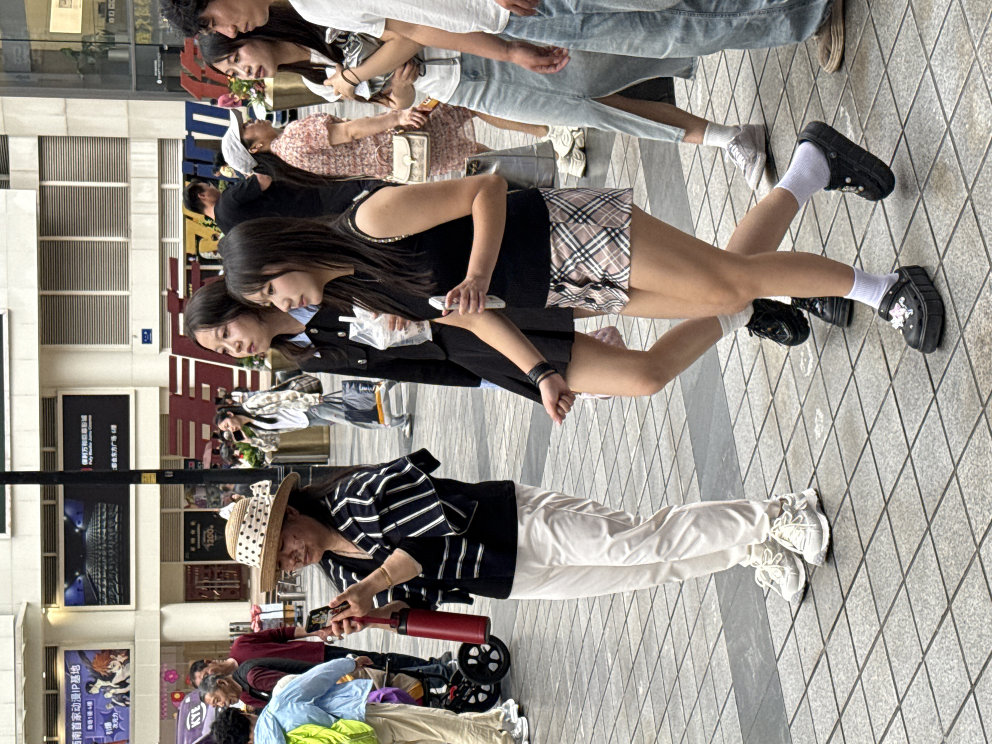 The image depicts an outdoor scene in the Yuzhong District of Chongqing, China, featuring a pedestrianized area with a diamond-patterned tile surface. Multiple people are present, including several young women, some of whom are looking at their phones. A woman in a striped shirt and a wide-brimmed hat holds a phone and what appears to be a red bottle or container. An elderly person in a wheelchair is visible in the lower left. A sign in Chinese mentioning "动漫IP基地" (Animation IP Base) and "都会东方广场6樓" (Metropolitan Oriental Plaza 6th Floor) is prominently displayed on a building in the background. Several posters are also visible, including one that mentions "保利万和巨幕影" (Poly Wanhe Giant Screen Film). The overall scene appears to be a busy urban area with people engaged in various activities.
