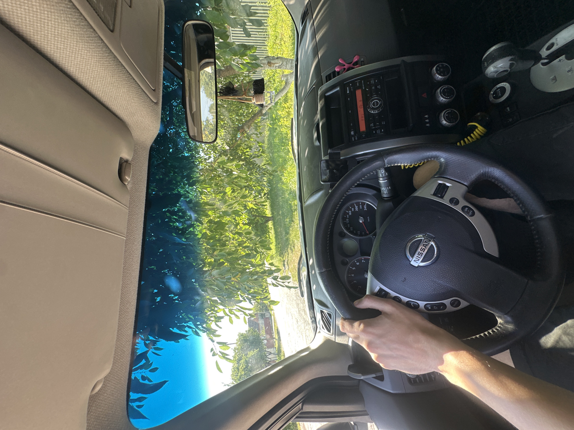 The image shows the interior of a Nissan SUV, viewed from the driver's perspective. A person's left hand is on the steering wheel. Through the windshield, a tree-lined street is visible with houses in the background. The location is identified as Shatava, Ukraine.