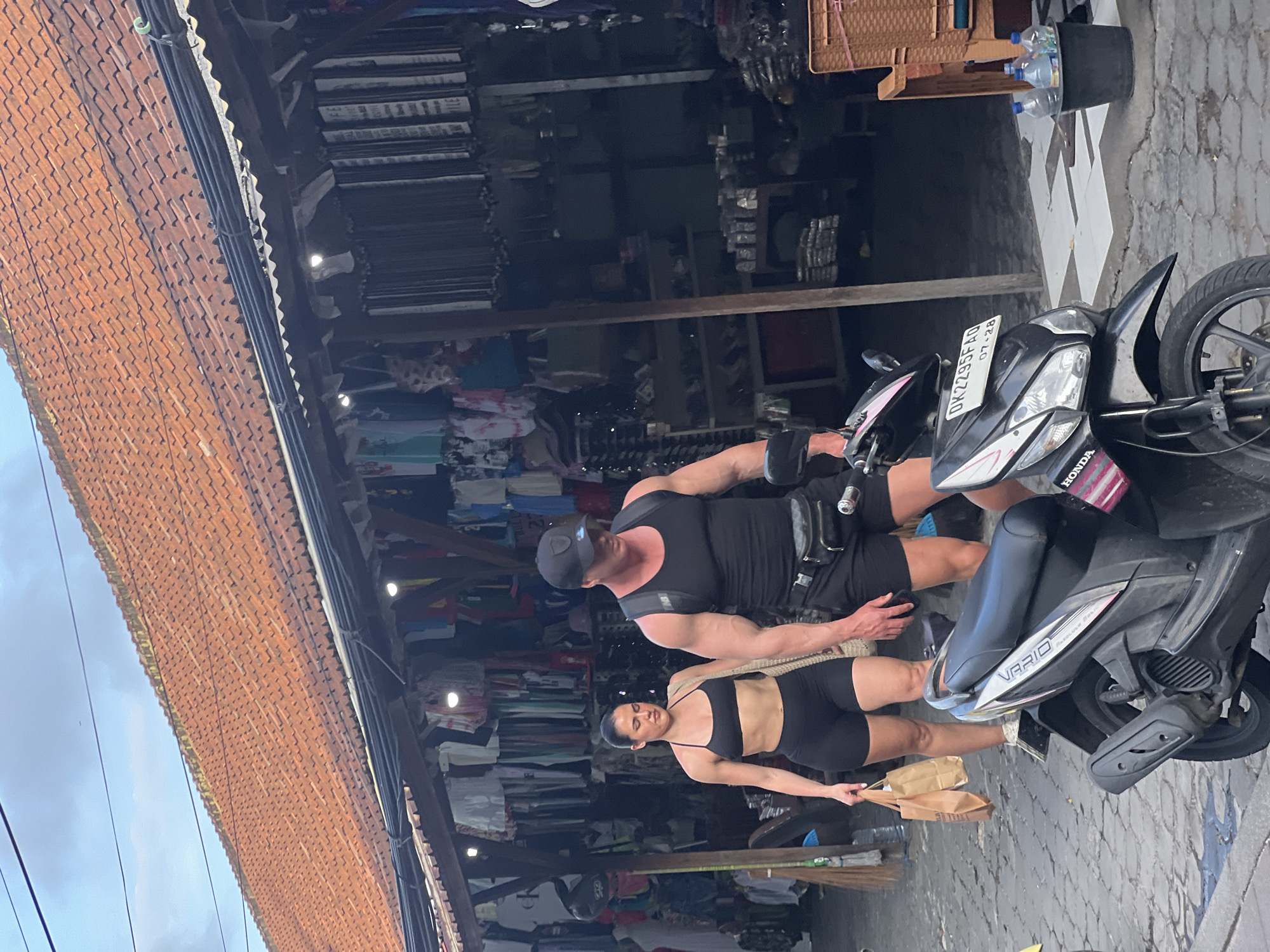 In Kuta, Indonesia, a muscular man in a black tank top, black shorts, and a black baseball cap walks alongside a woman wearing a black sports bra and black shorts. The man has a fanny pack around his waist and is leaning slightly forward. The woman carries two brown paper bags in her left hand. They are positioned in front of a market stall with an orange tiled roof, displaying various clothing items and goods. A black Honda Vario scooter is parked in the foreground, with its license plate reading "DK2295FAQ". In the background, more market stalls and a few bottles of water in a black bucket are visible. The ground is paved with cobblestones. The overall scene depicts a street market in a tropical urban environment.
