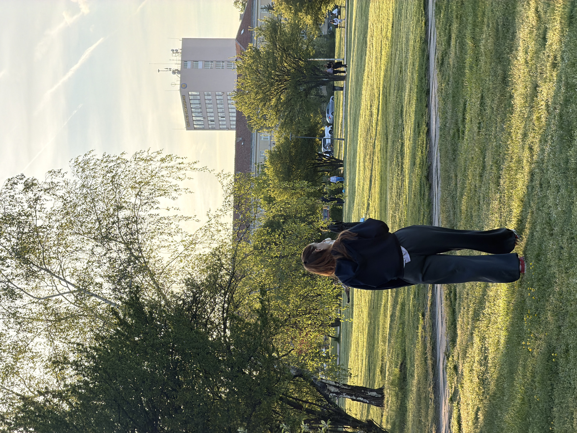 This image depicts a serene outdoor scene, likely a public park or a spacious green area, bathed in the warm, golden light of late afternoon or early evening. The setting is identified as Sofia, Bulgaria.
In the foreground, a woman with reddish-brown hair, dressed in a dark jacket over a white top and dark pants, stands with her back to the camera. She appears to be observing the landscape before her. A narrow dirt or gravel path winds through the vibrant green grass that dominates the mid-ground, showing patches of light and shadow from the low sun.
Various trees with lush green foliage are scattered across the scene, some closer to the viewer, others further back, providing a natural barrier between the park and the urban elements. In the background, a large, light-colored, multi-story building stands prominently. Its facade displays visible text: "МИНИСТЕРСТВО НА ЗЕМЕДЕЛИЕТО И ХРАНИТЕ," which translates from Bulgarian to "Ministry of Agriculture and Food," confirming the location in Sofia. The building features many windows and several antennas on its roof. Beyond it, hints of other structures with traditional red roofs are visible.
Several indistinct figures and parked cars can be seen in the mid-distance near the buildings, suggesting a public space frequented by people. The sky is bright and clear with a soft, yellowish hue, marked by faint contrails. The overall atmosphere is peaceful and contemplative, with the golden hour lighting enhancing the natural beauty of the park.