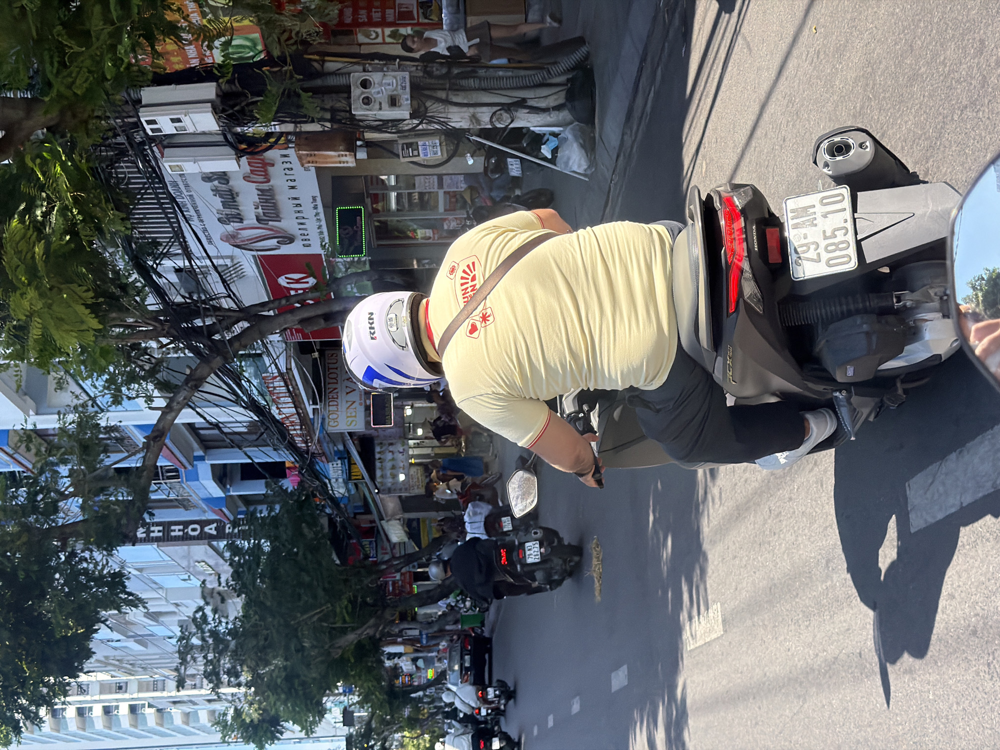 The image captures a street scene in Nha Trang Ward, Vietnam, likely during daylight. The perspective is from the viewpoint of someone riding a scooter. In the foreground, the rear of a dark-colored scooter is visible, with a license plate reading "29-AM 085.10". A person wearing a yellow t-shirt with a graphic design and a dark strap across their shoulder is seated on the scooter, their back mostly to the camera. They are wearing dark pants and white sneakers.
The street is paved with asphalt, marked with white lane lines. Several other scooters and a dark-colored car are visible on the road ahead, moving in the same direction. Pedestrians are present on the sidewalks, and buildings line the street. These buildings appear to be a mix of commercial establishments and residential structures, with balconies and signs. Trees with dense foliage are interspersed along the street, casting shadows on the road. Electrical wires are strung across poles and between buildings.
One building sign, partially obscured by trees, appears to be for a "GOLDEN LOTUS SEN VA". Another sign, also in the background, contains text in what appears to be Cyrillic script. The overall impression is of a bustling urban environment with active street life. The lighting suggests a sunny day, with strong shadows cast by the buildings and trees.