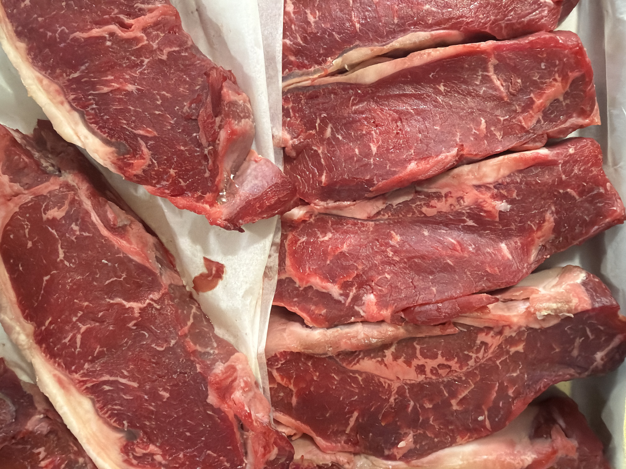 The image displays a close-up, overhead view of several raw beef steaks, likely on butcher paper. The steaks are a deep red color with visible marbling of white fat throughout. There are no people or any other objects in the frame. The background is white, suggesting a clean preparation surface. The steaks appear to be fresh and are arranged side-by-side. There is no indication of the specific location within Palmanova, Spain.