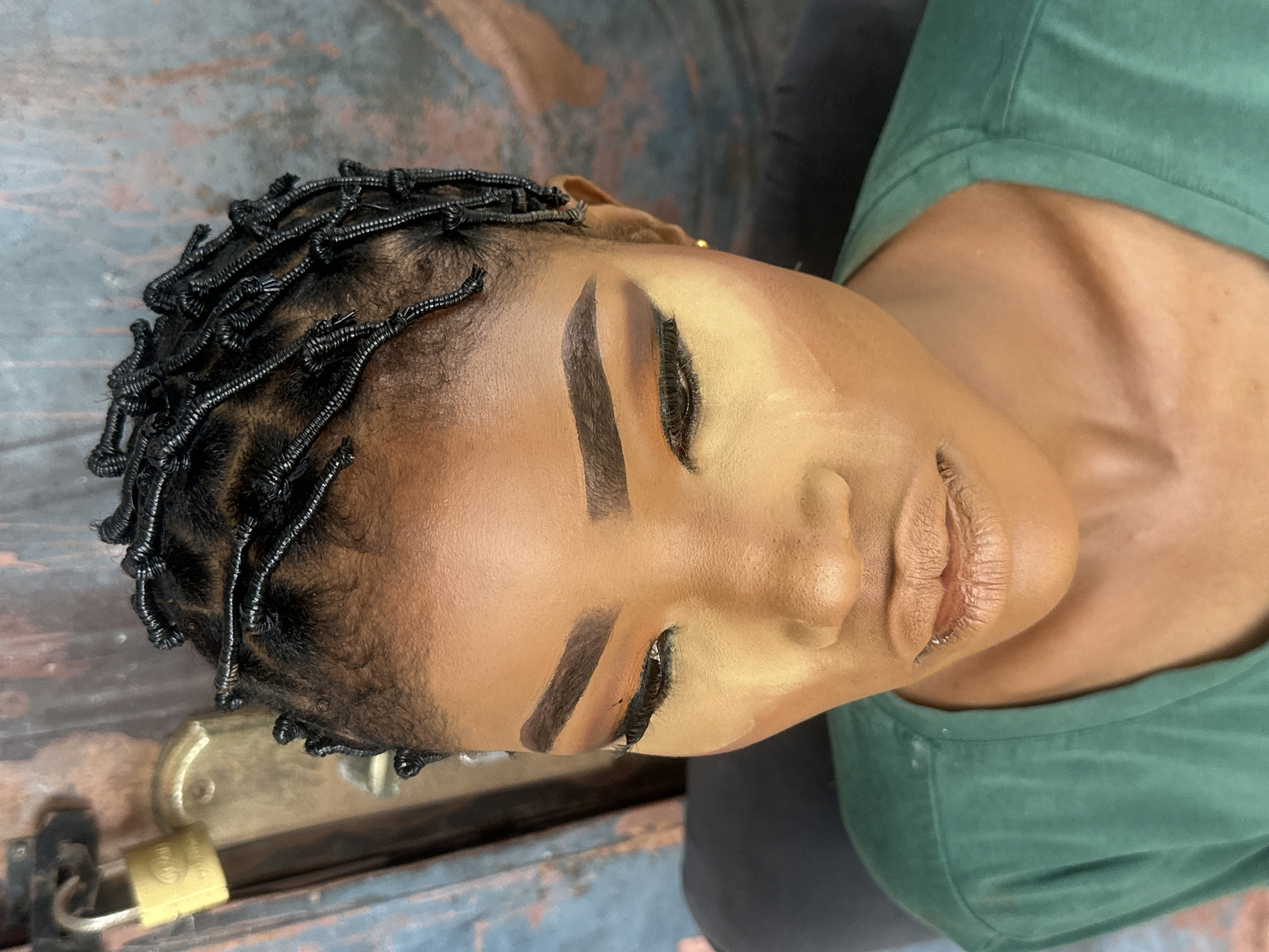 A close-up, eye-level shot captures a young Black woman's face from the upper chest to her tightly coiled black hair, with a padlock visible in the upper left corner. The woman is positioned slightly to the right of center, her head tilted down and to the left, and she appears to be looking down. She wears a dark green t-shirt.
Her skin is a rich brown hue, and she has dramatic eye makeup, including thick, dark eyebrows, warm-toned eyeshadow blending from a deep orange on the lid to a lighter tan in the crease, and a sharp black winged eyeliner. False eyelashes are applied. Her lips are coated in a matte nude lipstick. Her hair is styled into numerous small, tight coils, some of which are interwoven with black thread or string.
The background is a muted, textured surface with a vertical wooden element on the left side, where a yellow padlock is attached to a metal latch. The rest of the background consists of a dark, textured wall. A hint of a dark chair is visible in the lower left corner, and a portion of a dark shoulder is visible on the right.
The lighting is soft, with a slight shadow cast on the right side of her face, suggesting the light source is from the upper left.
The scene is an outdoor setting, likely a workshop or studio, given the utilitarian background and the focus on a makeup application. The padlock suggests a secure location, possibly a shop or a home with external storage. The presence of the padlock on a wooden structure, along with the textured wall, points towards a rustic or older building. This kind of setting is common in older parts of cities like Badagry, Nigeria, where traditional architecture and businesses often coexist.