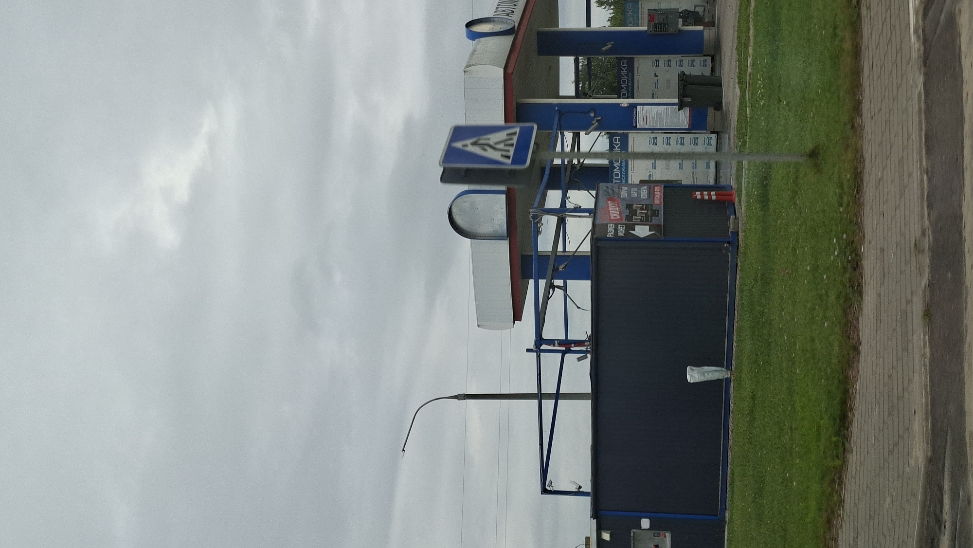 The image shows a view of a car wash facility in the city of Smalyavichy, Belarus. The car wash structure features blue pillars and a white roof with red trim. A pedestrian crossing sign is visible in the foreground, indicating a crosswalk. The car wash area appears to have multiple bays with blue metal framing. A dark blue modular building, possibly a service or equipment room, is situated to the left of the car wash structure. A trash can is visible near the car wash entrance. The ground is covered with grass and a paved walkway. The sky is overcast, with visible clouds. The overall scene depicts a typical urban or suburban commercial area.