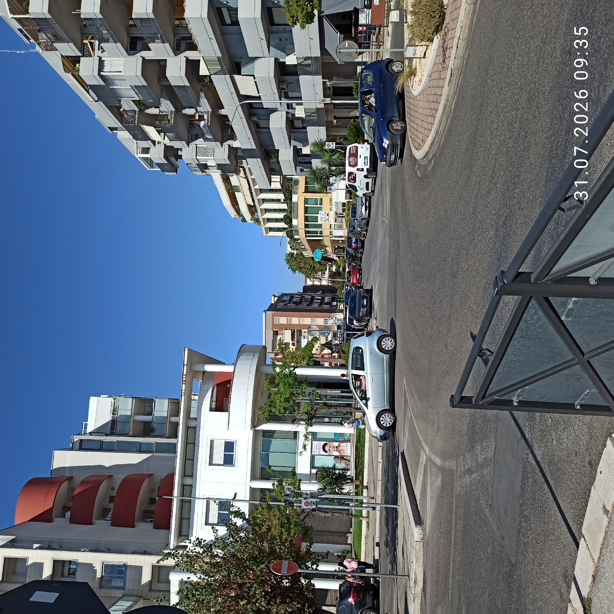 This is an outdoor, eye-level, medium shot photograph of a street intersection in Modugno, Italy, on a clear, sunny day. The image captures a multi-lane intersection with a mix of residential and commercial buildings lining the sides of the street.
On the left side of the intersection, a modern, multi-story building features a white facade with curved red balconies. Below this building, a lower white structure has large windows and columns. Trees and some shrubbery are visible in front of these buildings, along with a no-entry traffic sign. Several cars are parked or driving in the vicinity, including a dark-colored SUV. Two individuals are seen walking on the sidewalk.
The right side of the intersection is dominated by a large, grey, multi-story building with numerous balconies. This building appears to be an apartment complex or a mixed-use development. Below this building, commercial storefronts are visible, one with an awning. A blue Fiat 500 car is turning at the intersection, with a person driving it. Several other cars are in the intersection and on the road, indicating moderate traffic.
The road surface is asphalt, and there are street markings and a pedestrian crossing. A black metal fence with glass panels is in the foreground, partially obscuring the lower part of the scene. A timestamp in the bottom right corner reads "31.07.2026 09:35". The sky is a clear, vibrant blue. The overall impression is of a typical urban street scene in a Mediterranean city.