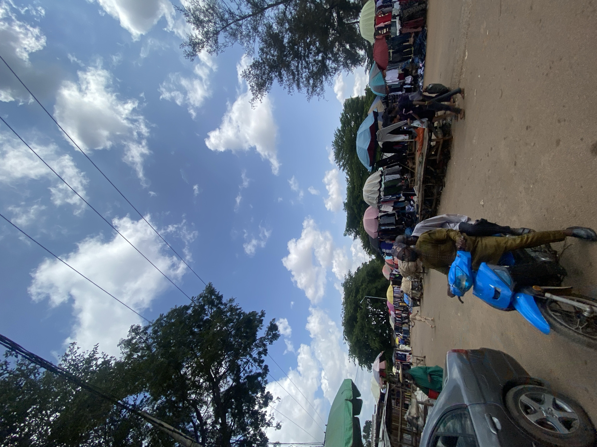 The image depicts an outdoor marketplace scene, possibly in Ado, Nigeria, under a partly cloudy sky. The foreground features a gray sedan parked on the left and a bright blue scooter with two men riding it. The man driving the scooter is wearing a brown outfit and a cap, while the passenger behind him is wearing a light-colored shirt. Behind the scooter, the market stretches out with numerous stalls covered by colorful umbrellas, displaying clothing and other goods. People are seen browsing and moving through the market. Trees and power lines are visible in the background, suggesting an urban or semi-urban setting. The ground is unpaved, indicating a dirt or dusty road surface.