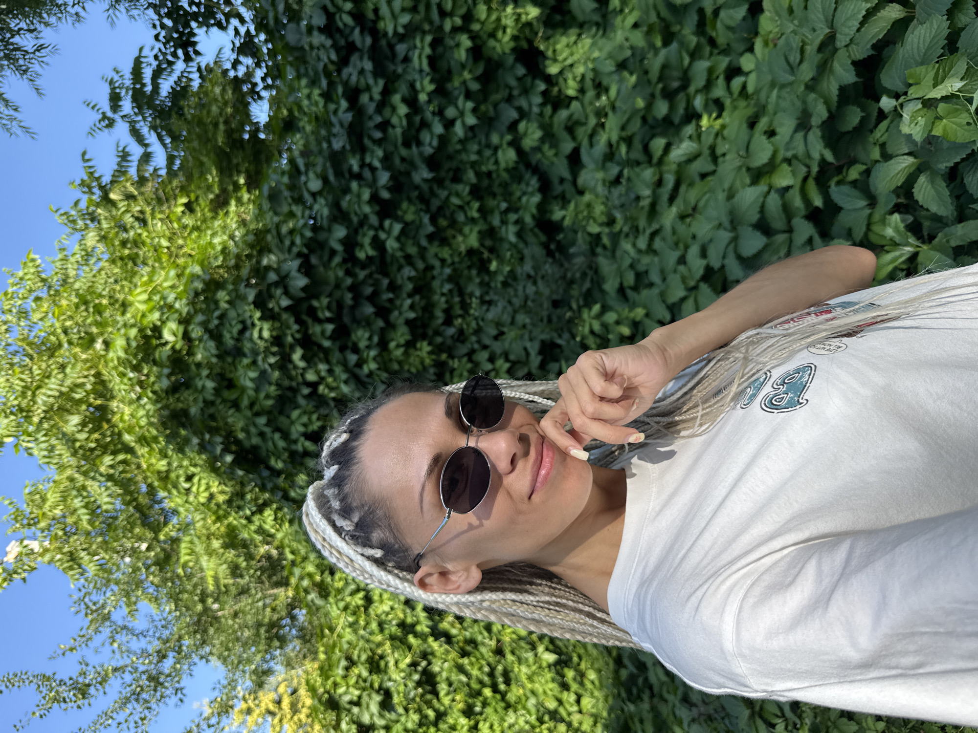 A woman with blonde braids and sunglasses is pictured in a selfie outdoors. She is wearing a white t-shirt and is standing in front of a wall of green foliage. The location is identified as Rusanovo, Russia. The lighting suggests it is daytime, and the sky is visible in the background. The woman's hand is near her face, with her fingers touching her chin. The image is focused on the woman, with the foliage serving as a backdrop.