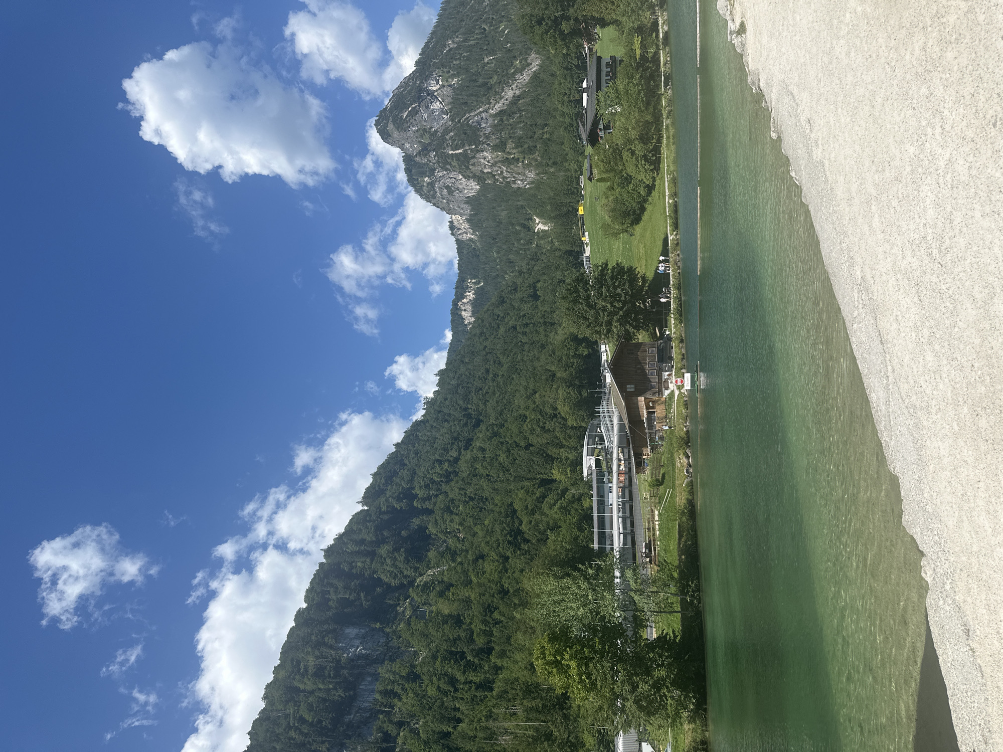 This is a vertical, eye-level, outdoor shot of a scenic lake and mountain landscape. The foreground is dominated by a sandy embankment that slopes down to the clear, turquoise water of the Königssee lake. The water is shallow near the shore, revealing the pebbled lakebed. The midground features the lake extending into the distance, with a small white boat visible in the water.
On the far bank of the lake, there is a green grassy area with scattered trees. Beyond the trees, several buildings are visible, including a large, modern-looking complex with white and gray structures, possibly a visitor center or recreational facility, and traditional wooden houses with dark roofs. Further up the slope, more buildings are nestled among the trees.
The background is dominated by a large, verdant mountain, densely covered with evergreen trees. The mountain's rocky peak is visible against a bright blue sky filled with scattered white, fluffy clouds. The overall impression is one of natural beauty and a recreational area. The location is Schönau am Königssee, Germany, given the iconic landscape. There are several people visible in small groups along the shore of the lake and near the buildings.