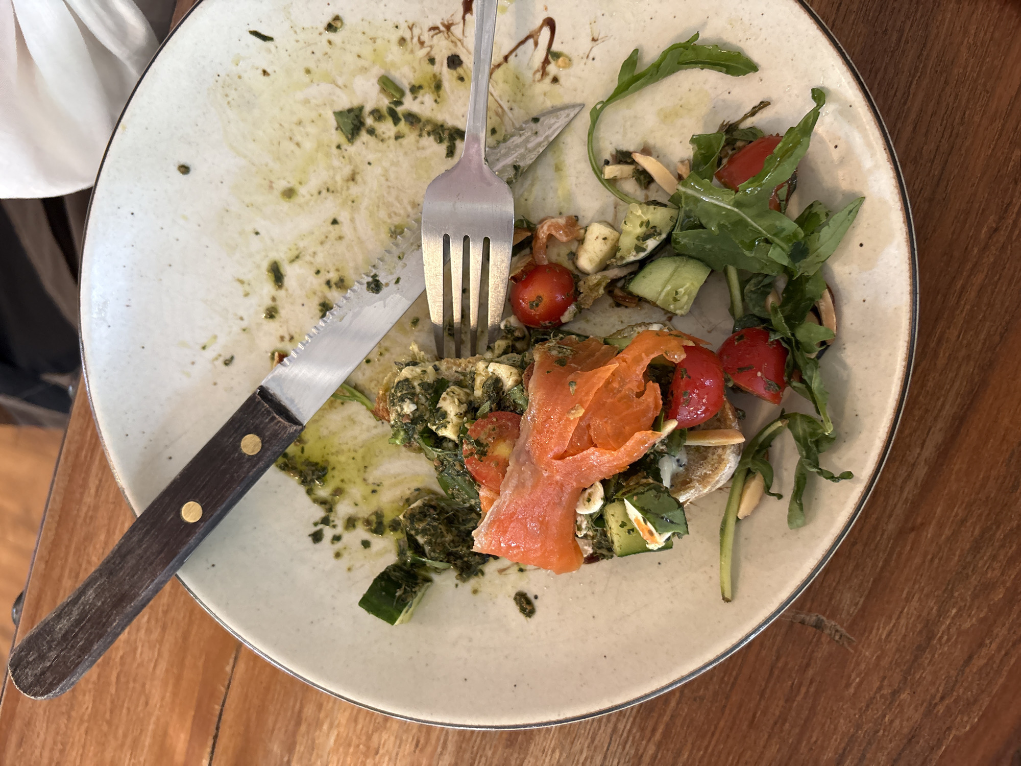 The image shows a close-up, overhead view of a plate with remnants of a meal, set on a wooden table. The plate, off-white with a thin dark rim, is mostly empty but displays scattered traces of pesto sauce, herbs, and some food items. These include sliced cucumbers, cherry tomatoes, and a piece of smoked salmon, still arranged on what appears to be a slice of bread or toast. A fork rests on the plate, its tines pointing towards the right, and a knife with a wooden handle is positioned to the left, its blade partially covering the plate's rim. The food remnants suggest the meal is either nearly finished or has been partially consumed. The overall scene is informal, likely a dining setting. The lighting suggests it could be daytime, possibly indoors or on a covered patio. There is no visible text in the image.
