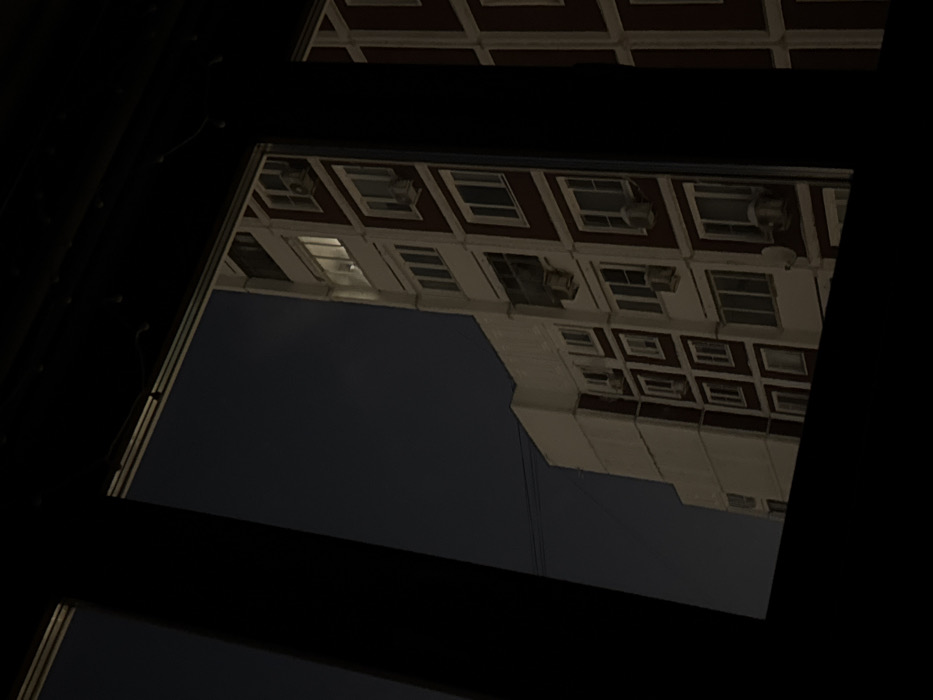 A low-angle shot frames a building with evenly spaced windows through a dark, gridded structure. The building’s façade, a mix of brick-red and white, features repeating patterns of windows and protruding air conditioning units. The sky is a dark, indistinct expanse filling the space around the building. The framing structure is black and appears to be metal, with rivet details visible. The overall image has a stark, somewhat industrial aesthetic.