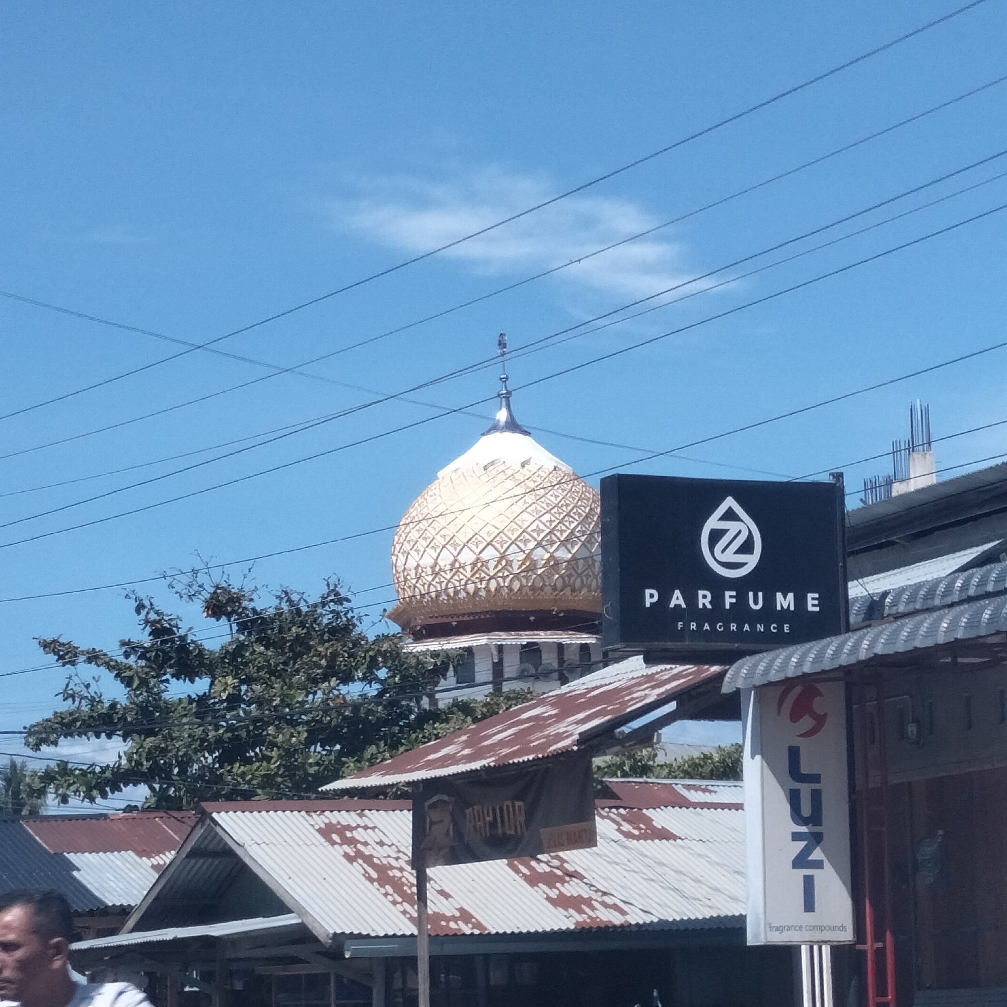 This outdoor, eye-level shot captures a street scene in Manetunong, Indonesia, under a clear, bright blue sky with a few wispy clouds. Numerous dark power lines crisscross the upper part of the frame.
The most prominent subject is the ornate, golden dome of a mosque, topped with a silver finial, which stands tall against the sky. Its surface is intricately patterned, and the base of the dome is white. In the foreground, various commercial buildings with weathered corrugated metal roofs, some showing patches of rust, line what appears to be a street. Green foliage and trees are visible, particularly around the mid-ground, partially obscuring other structures.
Several commercial signs are visible. A large black sign features a white "Z" logo with "PARFUME" and "FRAGRANCE" written below it in white. To its right, a vertical white sign displays "LUNI" in blue letters, with "fragrance compounds" at the bottom. Another banner, hanging beneath a corrugated roof, partially reads "RAPTOR". Construction rebar is visible on top of one building in the background, suggesting ongoing development.
In the bottom left corner, a partial view of a man's face, with short dark hair, is visible, looking towards the left out of the frame. The scene depicts a sunny day in a commercial or mixed-use area, characteristic of a town or city environment.