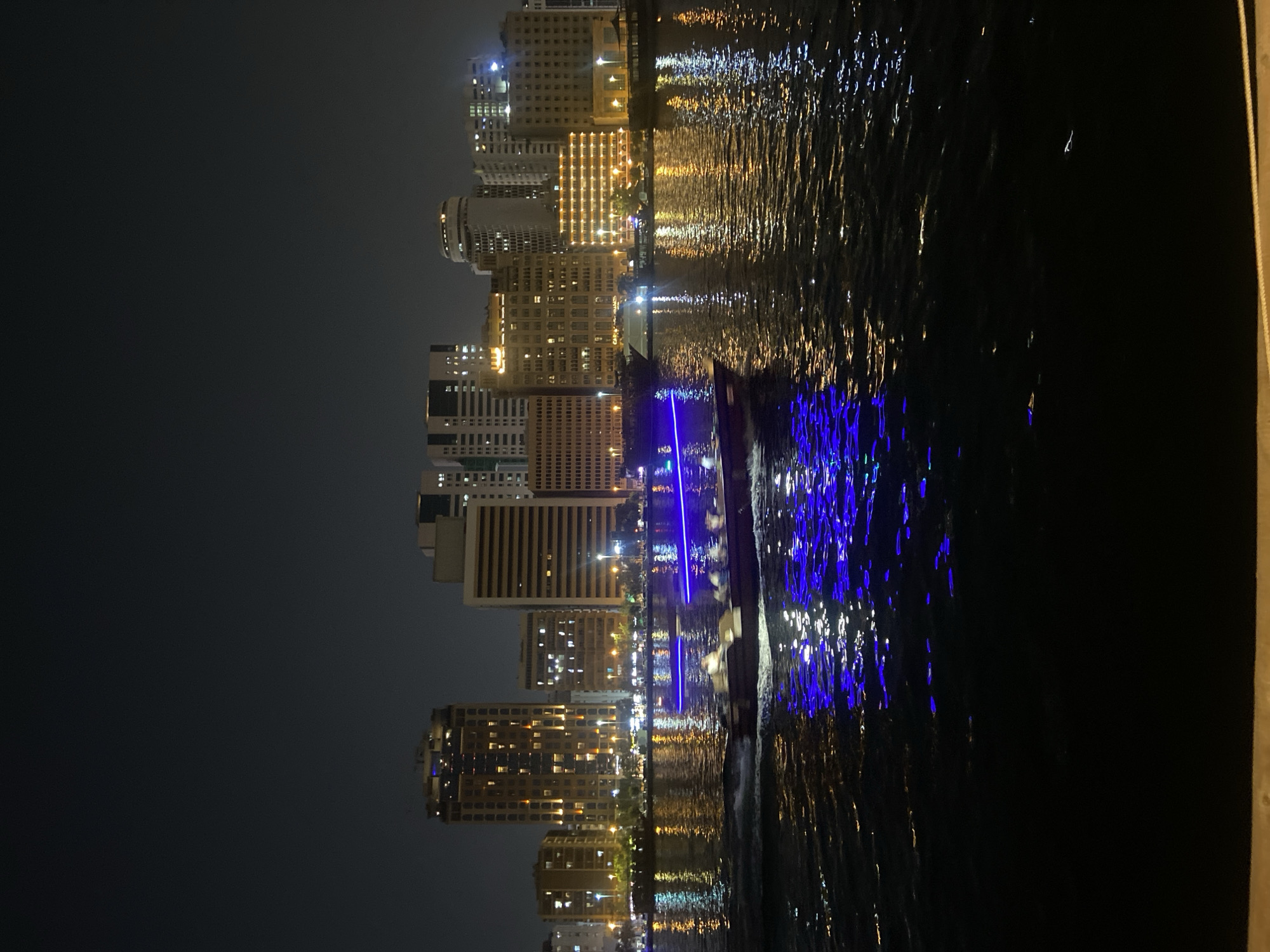 A low-angle, vertical shot captures a cityscape at night from across a body of water in Deira, United Arab Emirates. The dark sky looms above a densely packed skyline of illuminated buildings, ranging in height from mid-rise to skyscrapers. Many windows are lit, casting a warm glow from within. The water in the foreground reflects the vibrant lights of the city, creating streaks of orange, white, and electric blue on the dark surface. In the middle ground, a small boat with people on board navigates the water, leaving a trail of blue light from its undercarriage. The boat is moving from left to right across the frame. The overall scene is one of urban nightlife and activity against a dark backdrop.