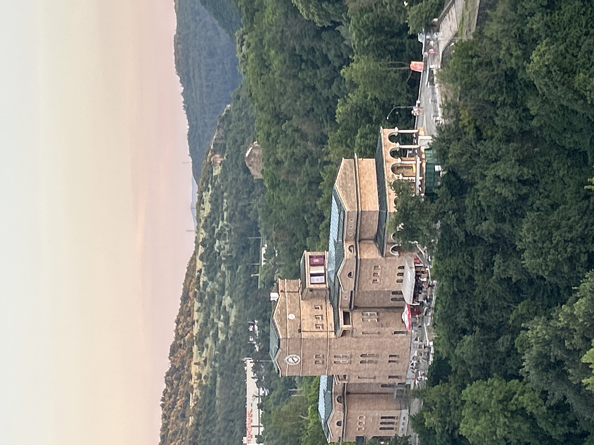 The image shows a panoramic view of a building in Veliko Tarnovo, Bulgaria, nestled amidst rolling, tree-covered hills. The building appears to be a historic structure, likely a cultural institution or a landmark, constructed with light-colored brick. It features a prominent clock tower and arched colonnades. In front of the building, there is an outdoor seating area with tables and umbrellas, suggesting a cafe or restaurant. A few people are visible in this area, engaged in conversation or dining. The surrounding landscape is lush with green foliage, with hints of other structures visible in the distance on the hillsides. The sky above is a soft gradient of pale pink and orange, indicating either dawn or dusk. The overall scene conveys a sense of tranquility and historical charm.