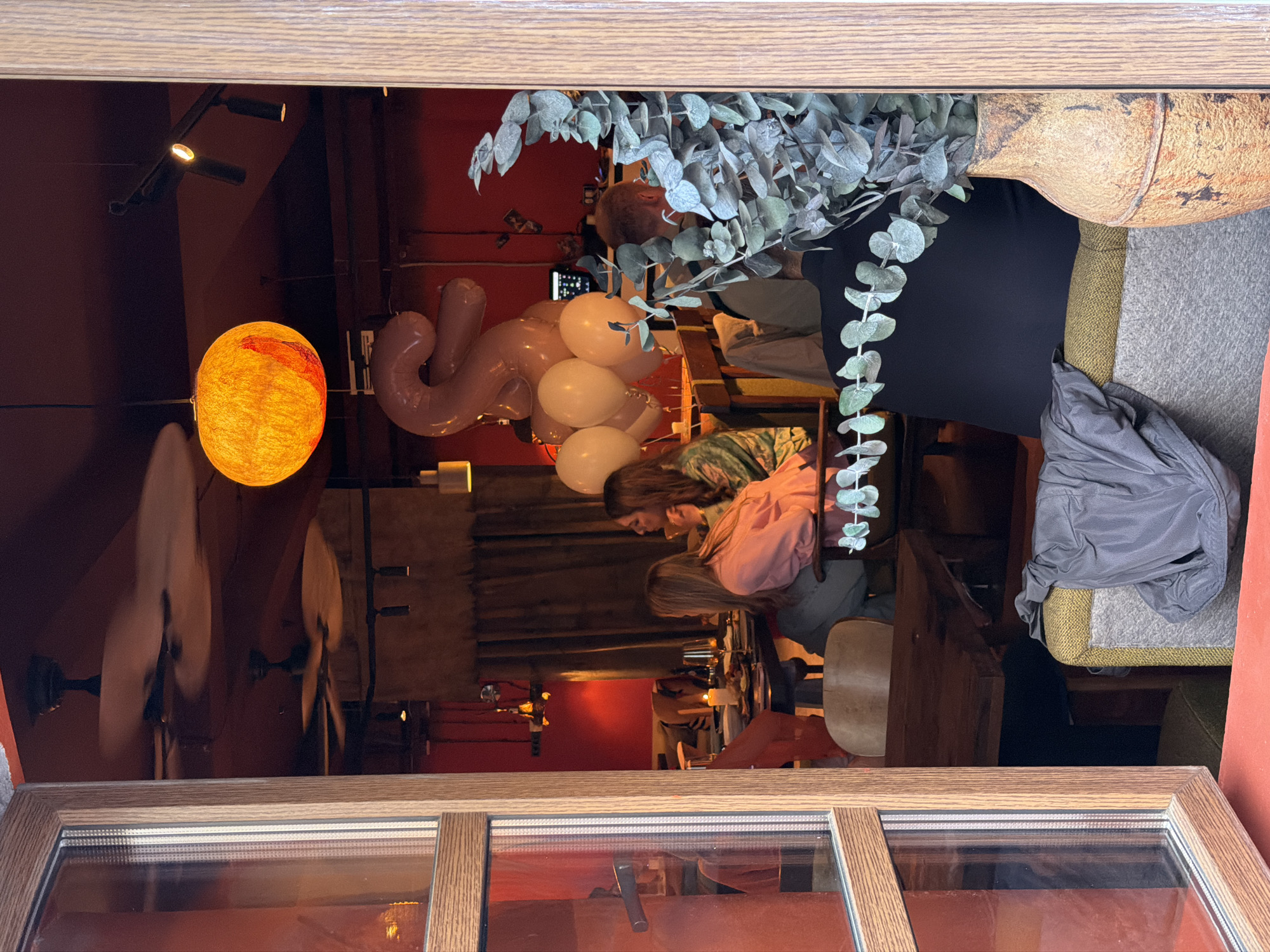 A slightly ajar window frames an interior view of a restaurant or bar. The location is potentially in Moscow, Russia. A spherical orange light fixture and pink letter-shaped balloons are visible, suggesting a celebratory atmosphere. Two women are seated at a wooden table. A decorative eucalyptus arrangement sits on a table or surface nearby. The background features red walls and other restaurant patrons. A rotating ceiling fan is visible in the reflection of the window, implying indoor air circulation. The scene is captured from an outside perspective looking into the establishment.
