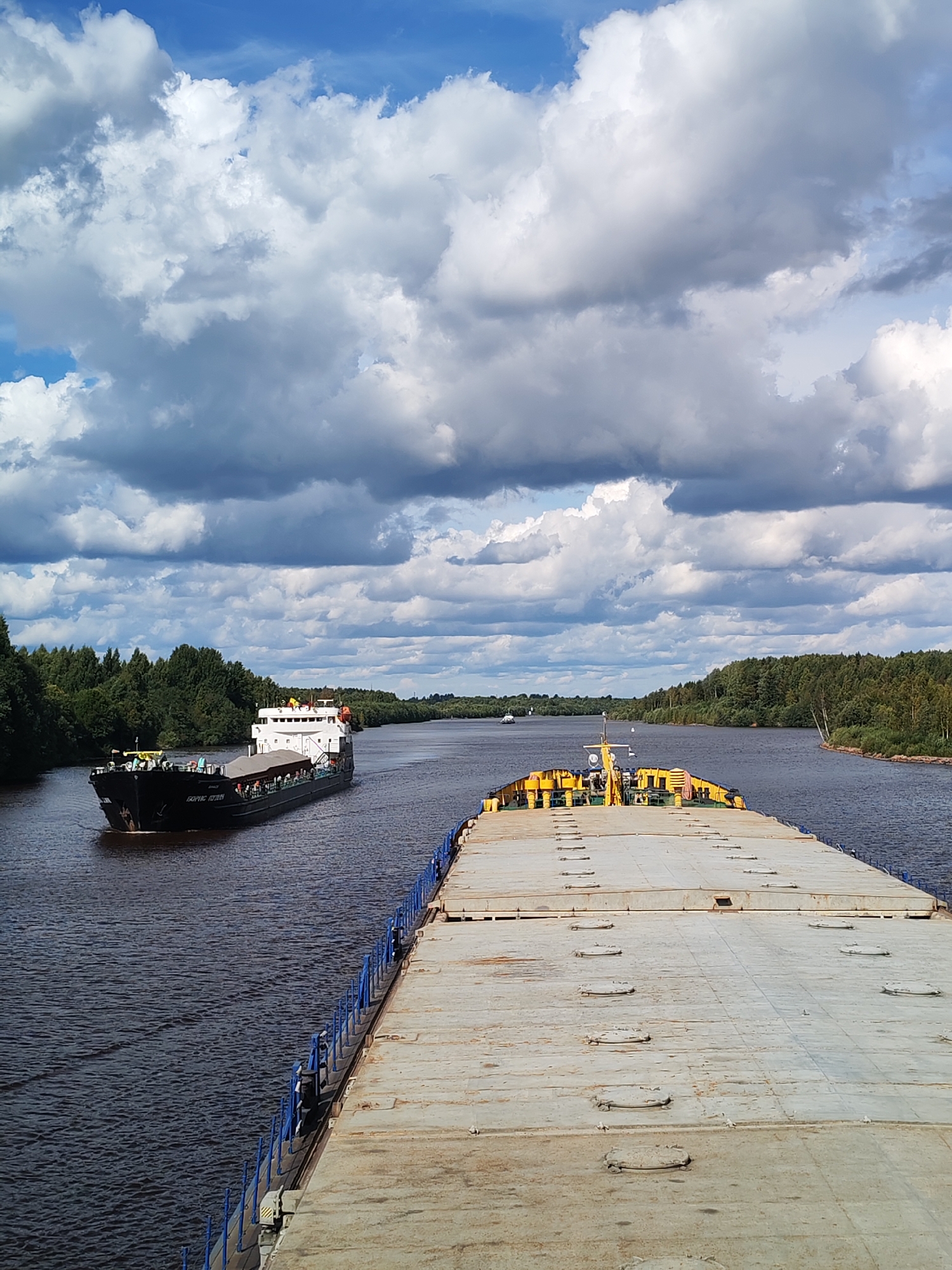 The image depicts a view from the deck of a vessel, looking forward down a wide river. On the left side of the river, a black and white tanker ship is visible, proceeding in the same direction. The tanker has a white superstructure with visible windows and some deck equipment. The bow of the tanker displays Cyrillic lettering that appears to read "БОРИС ЩЕРБИН". The river is flanked by dense, green trees on both banks, forming a natural landscape. The sky above is filled with dramatic, volumetric clouds, casting shadows on the water and landscape, suggesting dynamic weather. In the foreground, the deck of the current vessel is prominent, made of weathered, light-colored material with numerous circular hatches. A blue railing runs along the edge of this foreground vessel. In the distance, another vessel is faintly visible on the river, and the river curves to the right, disappearing behind the tree line. The scene is consistent with a navigable waterway in a rural or natural setting. The text overlay confirms the location as Annensky Most, Russia.