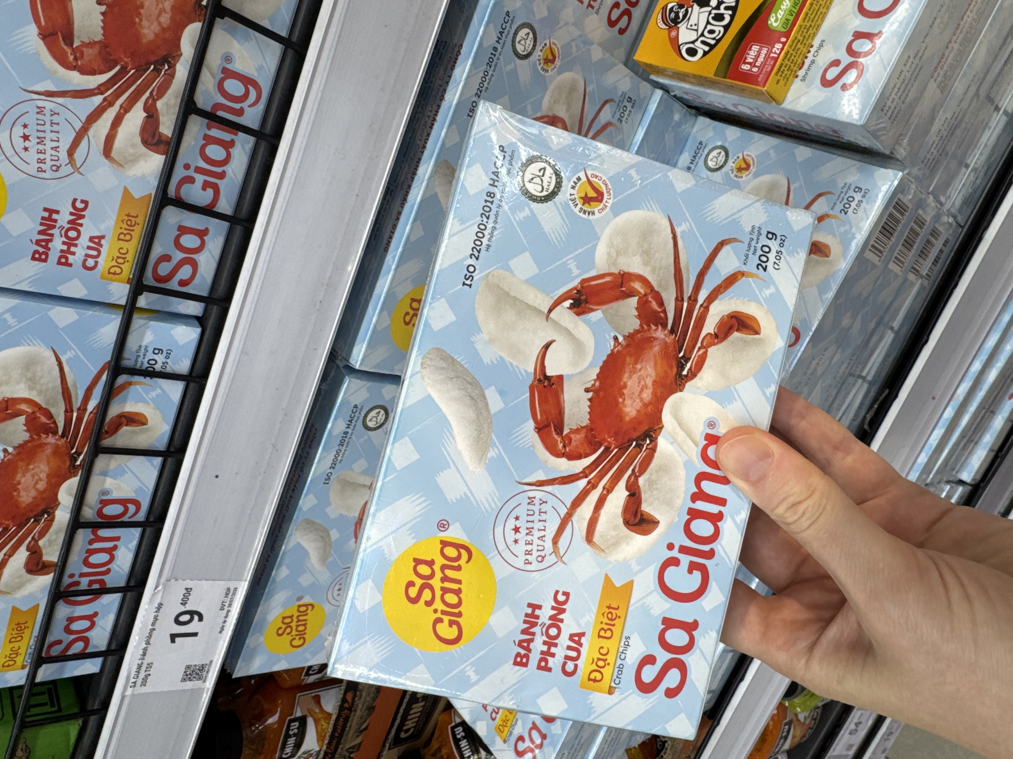 The image shows a close-up of a hand holding a box of "Sa Giang" brand crab chips in a supermarket aisle in Da Nang, Vietnam. The box is light blue with a graphic of a red crab and white puffed snacks. The text on the box is in Vietnamese and English, indicating "Bánh Phồng Cua" (Crab Puffs) and "Đặc Biệt" (Special). There are also labels for "Premium Quality," "ISO 22000:2018 HACCP," and "Halal." In the background, more boxes of the same product and other food items are visible on shelves. A price tag in Vietnamese currency (đồng) is partially visible on the shelf in the upper left corner. The environment appears to be a brightly lit, organized retail space.