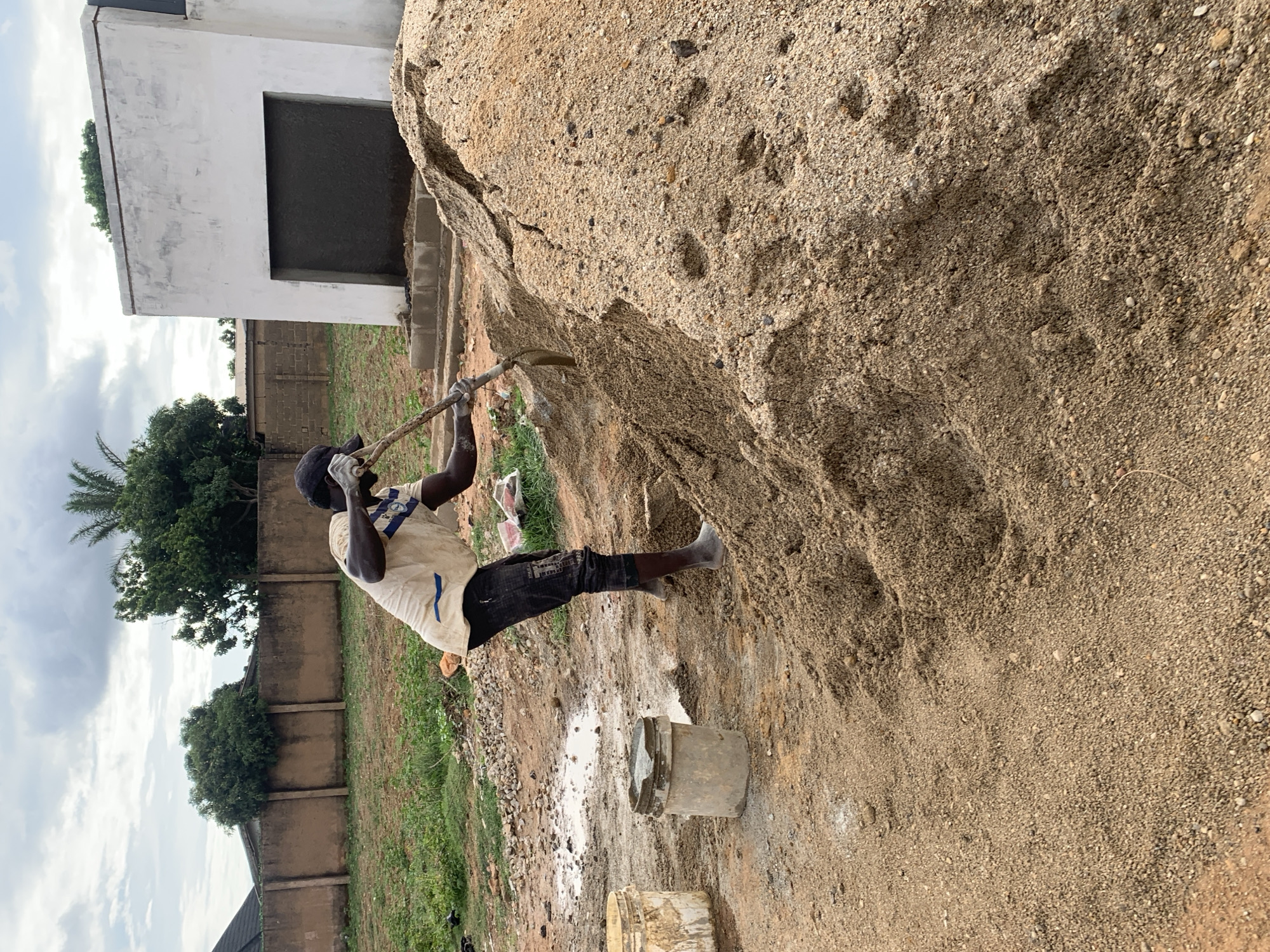 The image displays an outdoor scene in Jos, Nigeria, centered on a male laborer engaged in excavation. The man, with dark skin, is wearing a light-colored t-shirt with blue stripes, dark pants, a dark baseball cap, and white work gloves. He is positioned with his torso bent forward, actively shoveling light brown, coarse sand mixed with small pebbles from a large mound that occupies the right foreground. To his immediate left, two utilitarian plastic buckets, one light yellow and one grey, rest on the uneven ground near a muddy puddle and scattered gravel. The ground is a reddish-brown soil, unpaved and showing signs of disturbance. In the mid-ground, a tan-colored concrete block wall forms a boundary, with various green trees visible above and behind it. Further to the right in the background, a white, unadorned building with a prominent dark, rectangular recessed opening stands. The sky is overcast with grey clouds.