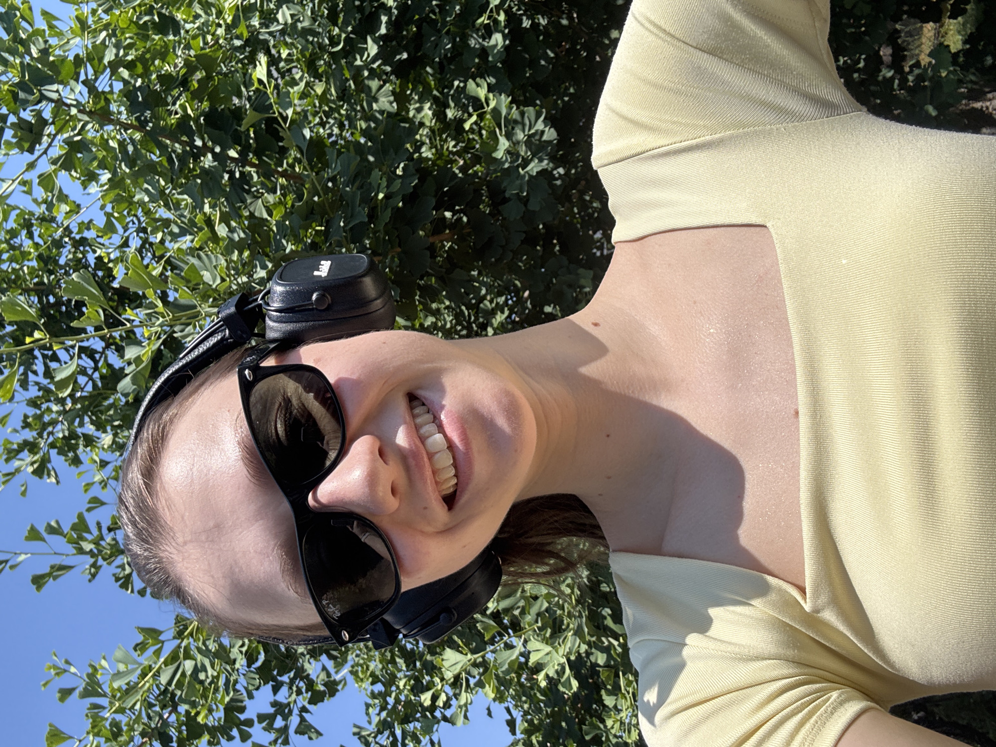 A young woman with brown hair is taking a selfie outdoors, with trees and a blue sky in the background. She is wearing a pale yellow, square-neck, ribbed top, black sunglasses, and black headphones. She is smiling and her teeth are visible. The scene appears to be in Wörth am Main, Germany, likely during the daytime given the bright sunlight and clear sky.