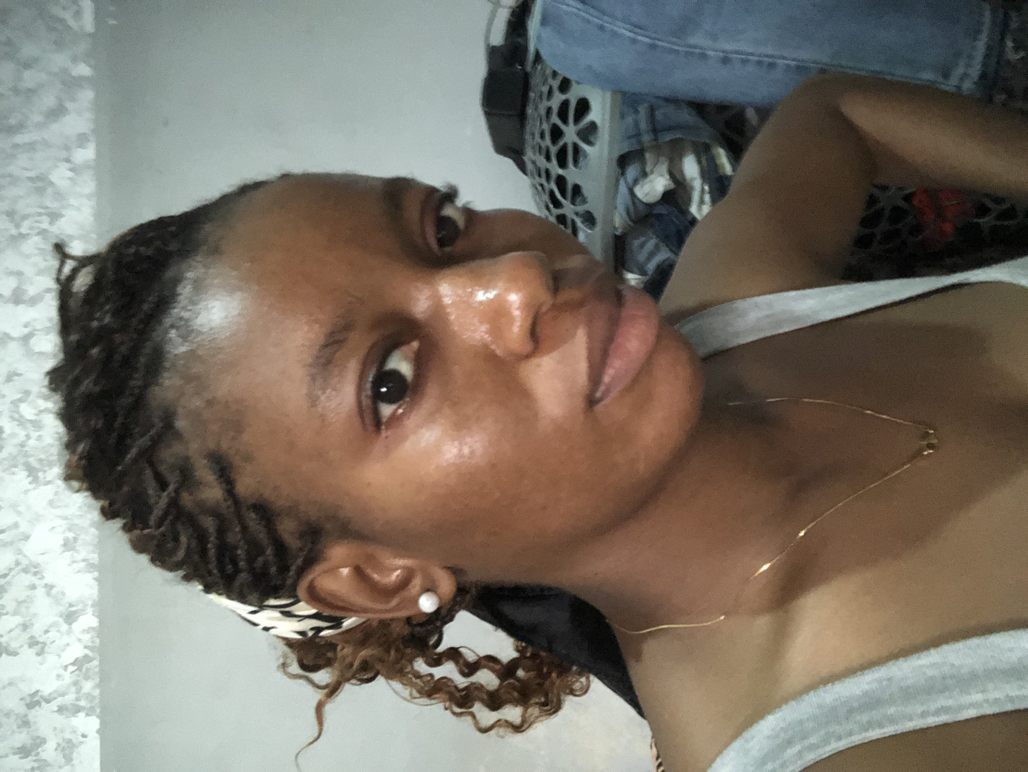 A close-up, vertically oriented photograph shows a Black woman from the chest up. She is reclining, wearing a light gray tank top and a delicate gold necklace. Her hair is styled in small braids with curly ends near her face, secured with a black and white patterned headband. A single pearl earring adorns her left ear. Her skin appears slightly shiny. Behind her, out of focus, is a blue laundry basket and some folded denim clothing. The background is a pale gray wall. The location is not specified. The city is not specified.