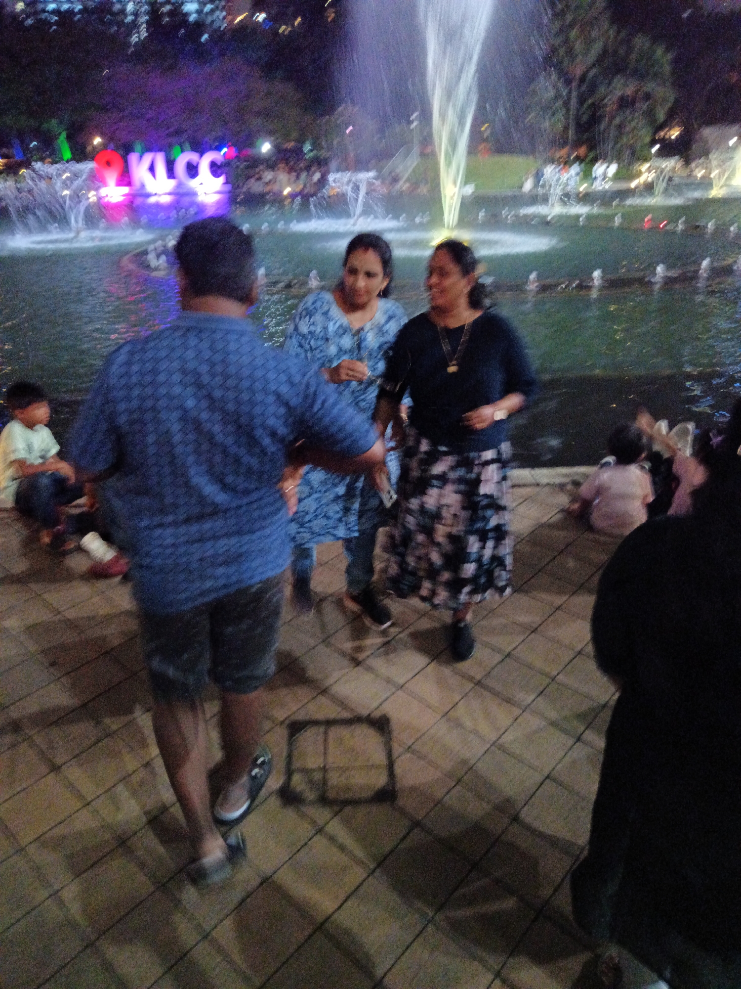 This is a nighttime, slightly blurred photograph taken at the KLCC Park in Kuala Lumpur, Malaysia. The foreground features the backs of several people, including a man in a blue patterned shirt and shorts, and two women. One woman wears a blue patterned top, and the other a dark long-sleeved shirt and a floral skirt. The background shows a water fountain with illuminated water jets and a sign that spells out "KLCC" in large, brightly lit letters. Other people are visible in the background, some sitting and some standing near the fountains. The ground is paved with square tiles.