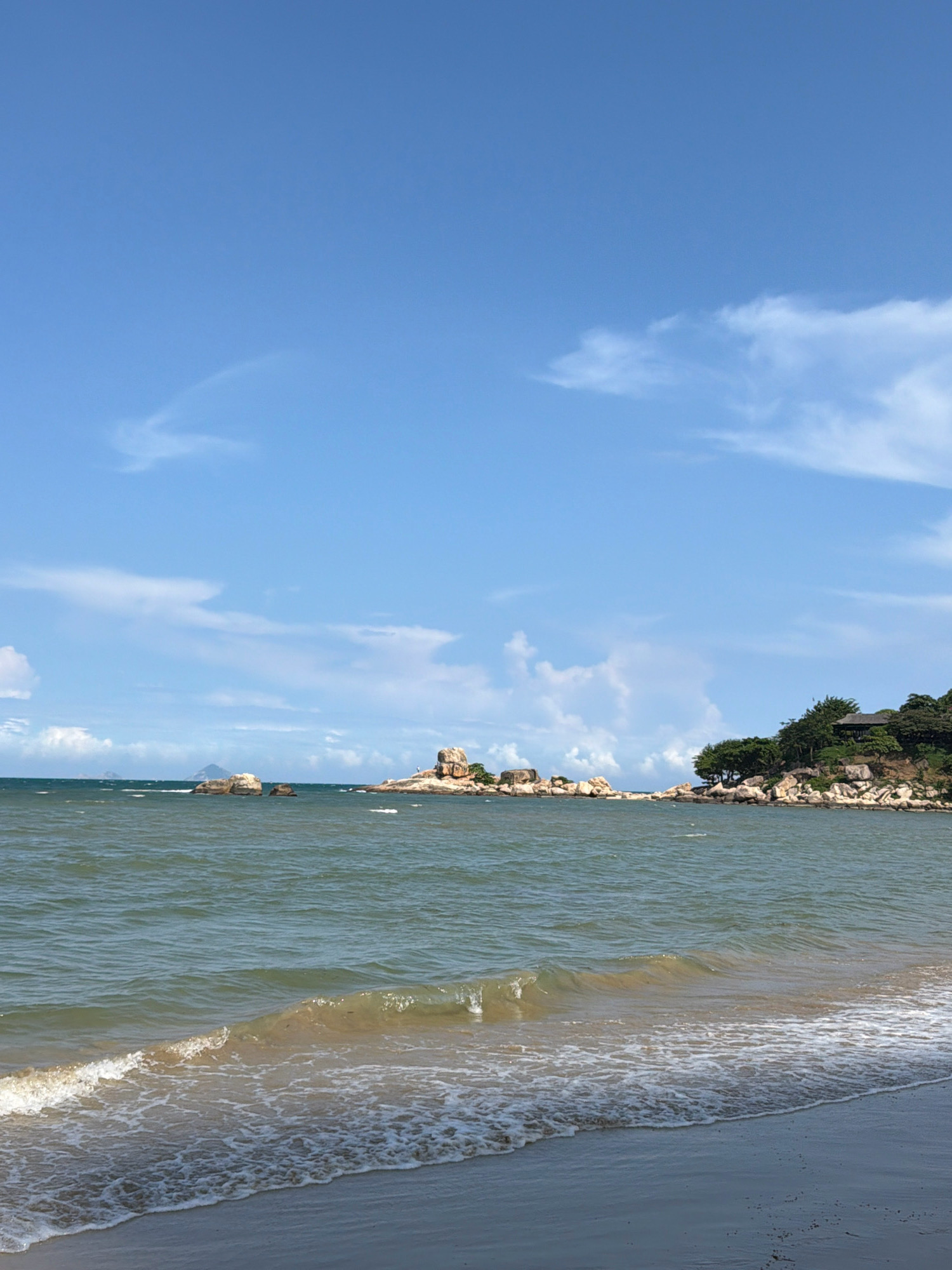 The image depicts a serene coastal landscape with a bright blue sky dotted with sparse white clouds. The foreground features a sandy beach with gentle waves lapping at the shore. The water is a greenish-blue hue, showing some choppiness. In the middle ground, several rocky formations rise from the sea, some of them quite large and prominent. One cluster of rocks in the distance appears to have a distinct, rounded shape.
To the right of the frame, a lush green hillside slopes down towards the water, covered with dense trees and shrubs. Perched on this hillside is a small, dark-roofed structure, possibly a pavilion or a small building, partially obscured by the foliage. The coastline extends to the left, revealing more rocky outcrops and the vast expanse of the ocean. The overall impression is one of a tranquil, natural coastal environment, likely a popular scenic spot. This scene is characteristic of areas around Nha Trang, Vietnam, known for its beautiful beaches and offshore islands with similar rock formations and vegetation.