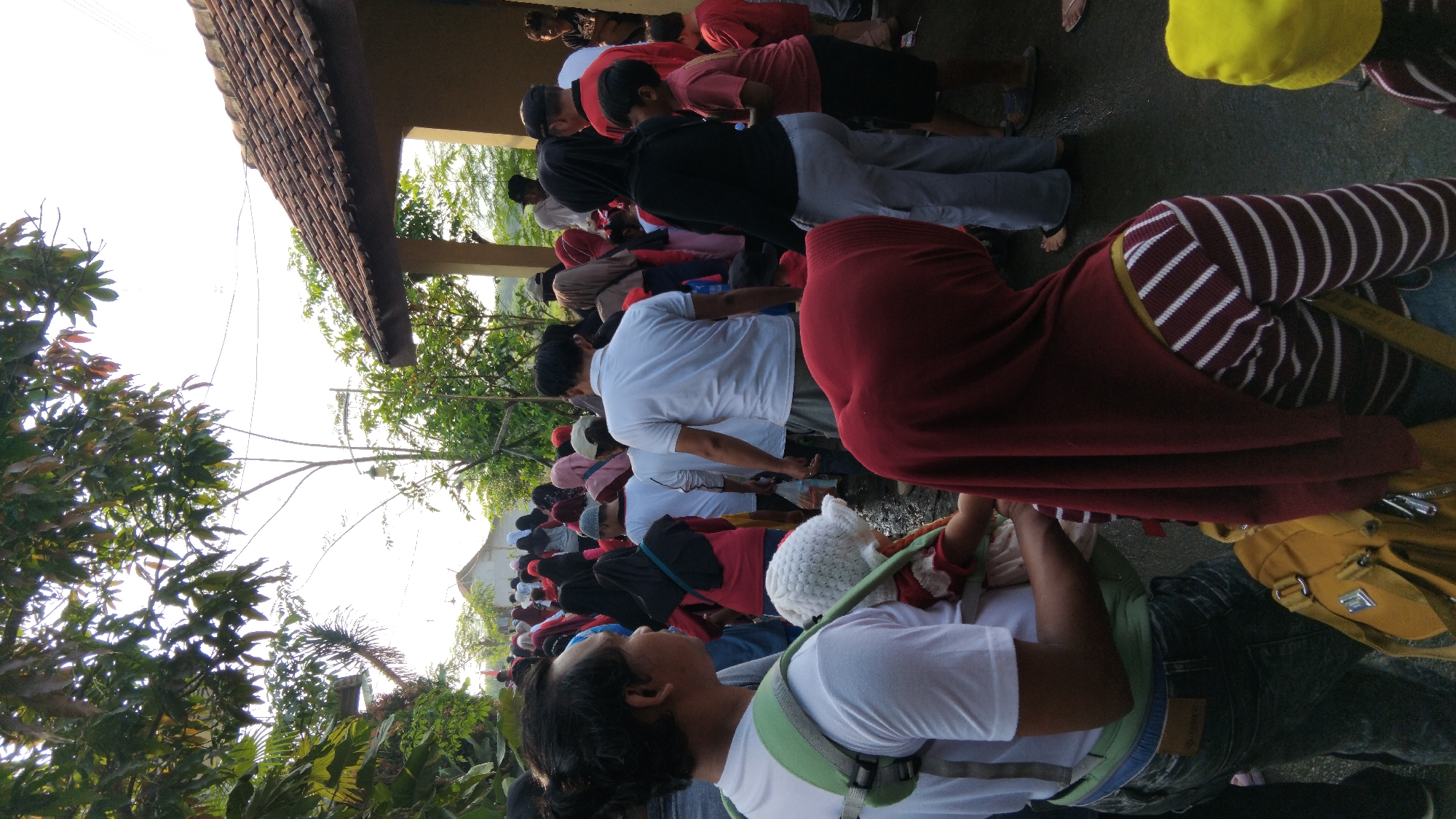 In a street scene in Posong, Indonesia, a crowd of people, mostly women and children, are gathered. The setting appears to be an outdoor event or gathering, with trees and a building with a tiled roof visible. Many individuals are wearing headscarves, and some are carrying children. A man in the foreground is wearing a white shirt and a baby carrier with a child in it. A large yellow bag is also visible in the foreground. The ground is wet, suggesting recent rain. The atmosphere seems to be one of community engagement and activity.