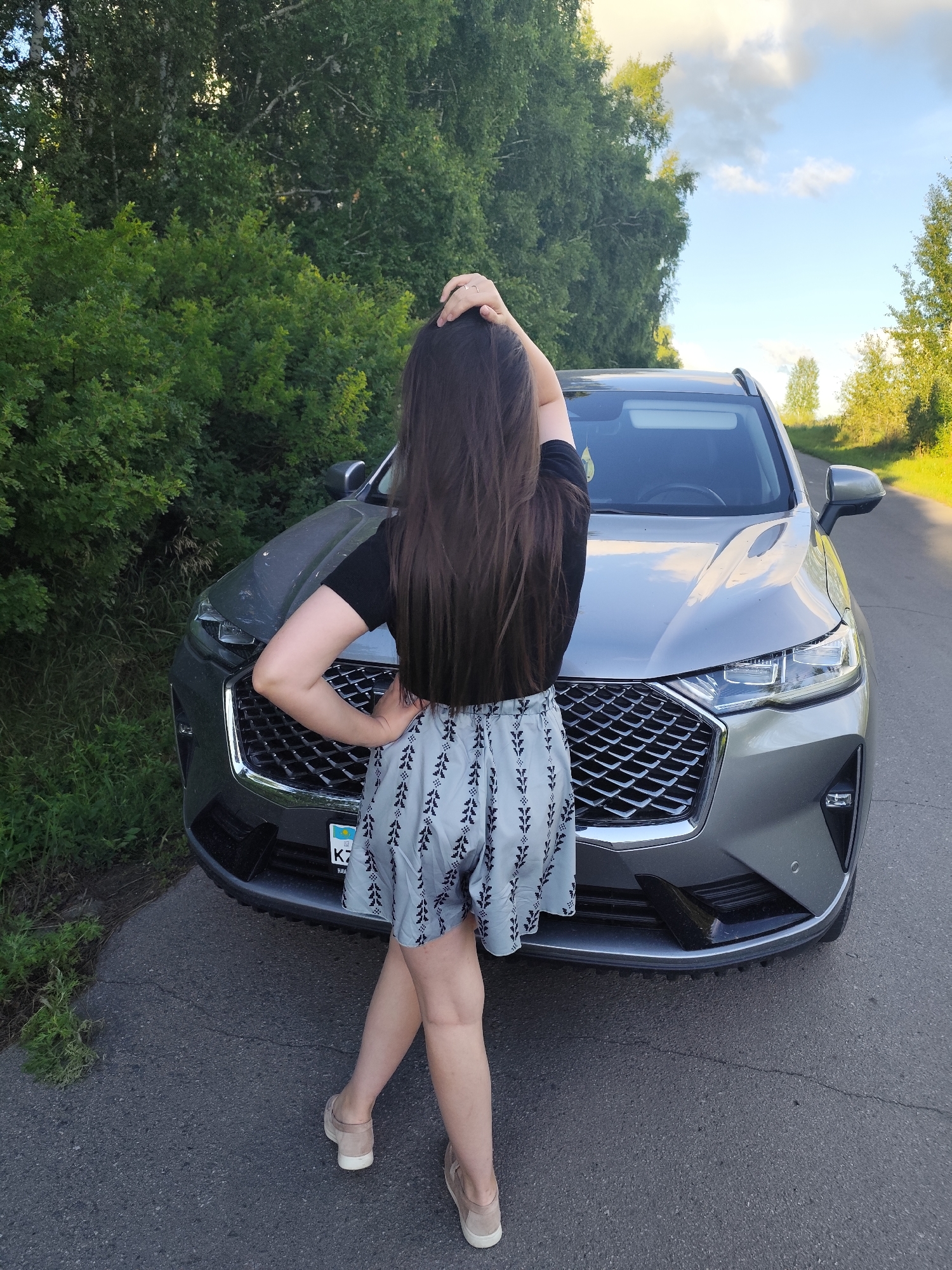 A woman stands with her back to the camera, facing a grey SUV parked on a paved road. She has long, dark hair, which she is holding with one hand while the other rests on her hip. She is wearing a black t-shirt and light-colored shorts with a dark leaf pattern. Her feet are clad in light beige slip-on shoes.
The SUV is a modern vehicle with a prominent grille and sharp headlights. A license plate with the letters "KZ" is visible on the front of the car. The background consists of lush green trees and foliage on either side of the road, with a cloudy sky overhead. The sunlight suggests it is late afternoon or early evening. The scene appears to be in a rural or semi-rural area, possibly a roadside.