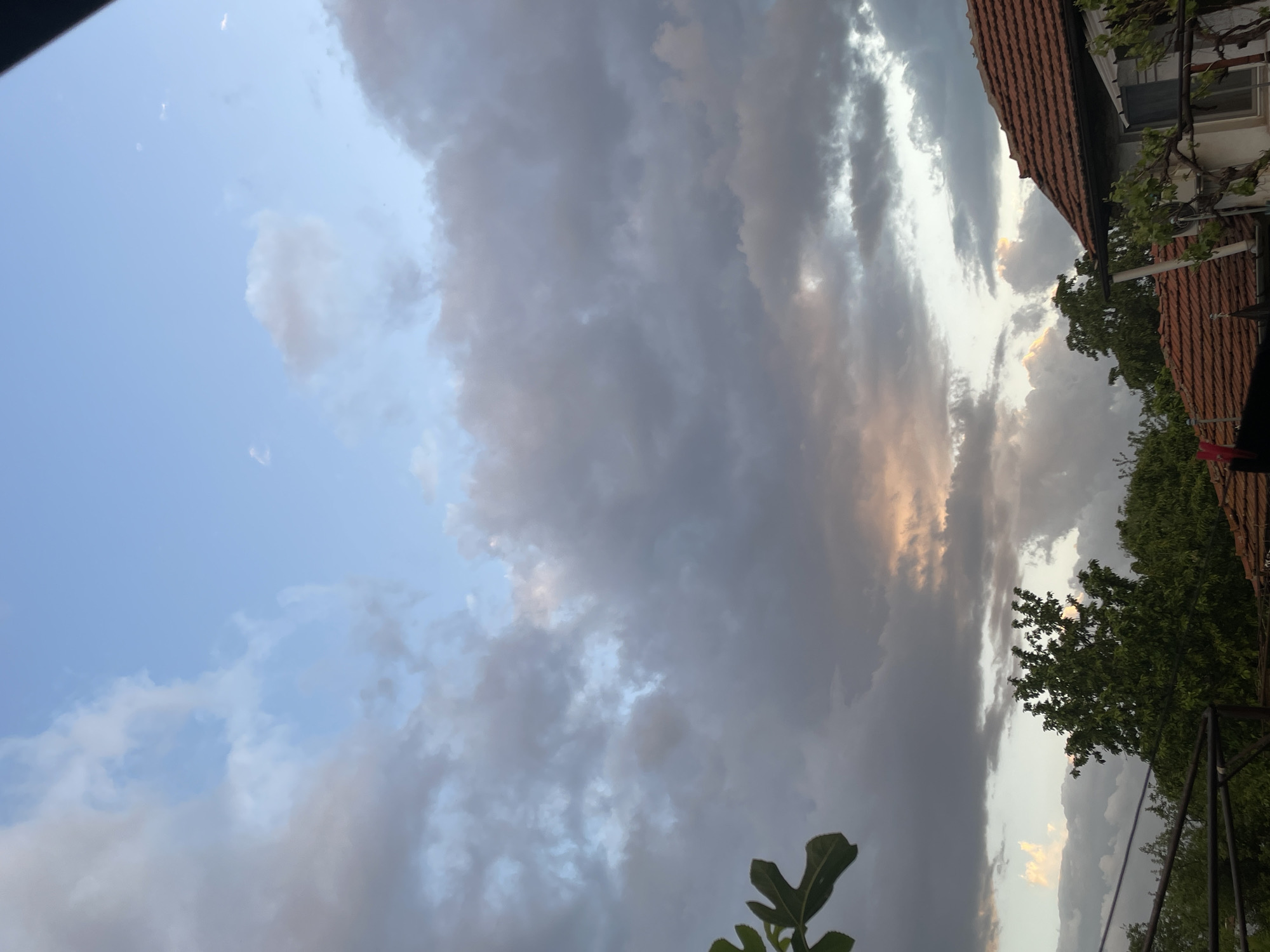 The image depicts a sky with a mix of blue and grey clouds, suggesting an overcast or potentially stormy weather condition. The view is from a ground-level perspective, looking upwards, with parts of a building, including a red tiled roof, and greenery like trees and plants visible along the right side of the frame. The presence of these elements suggests the image was taken in a residential area. Given the inclusion of a traditional red-tiled roof, this is consistent with the architectural styles seen in Sofia, Bulgaria, it is highly likely that the photo was taken in Sofia. The orientation of the image is rotated 90 degrees counter-clockwise.