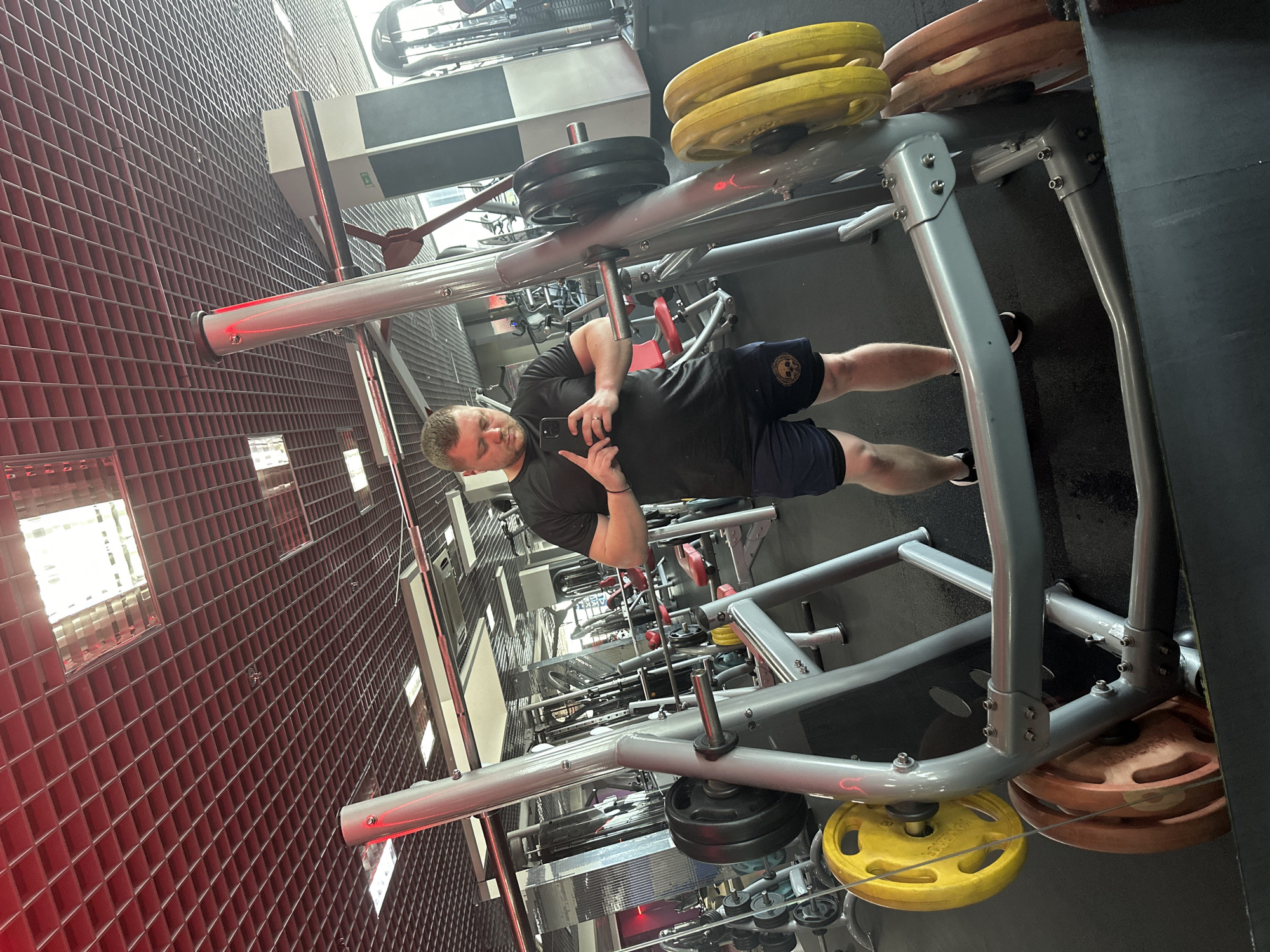 A man in athletic wear stands in a gym, taking a mirror selfie. He is wearing a black t-shirt and navy blue shorts with a skull emblem. He is holding a smartphone in his right hand. In the background, various gym equipment is visible, including weight racks with yellow and brown weight plates, a squat rack, and weight machines. The gym has a modern design with a red, tiled ceiling and bright lighting. The scene appears to be indoors, in a well-lit gym. There is no indication of the specific city or country.