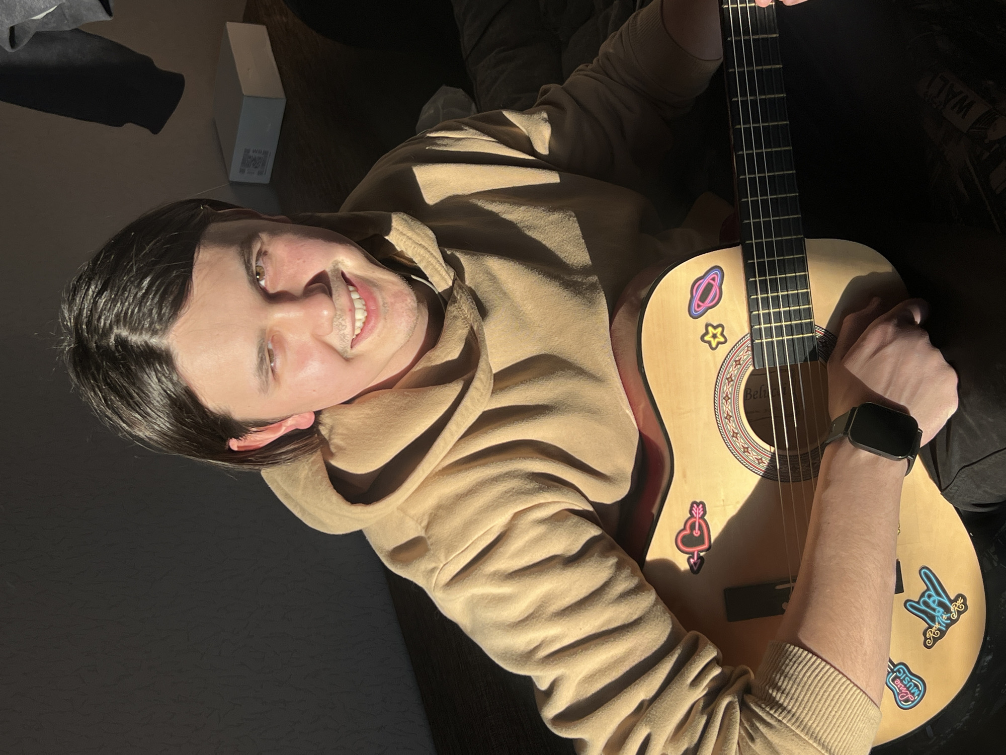 The scene, located indoors in Chaikovsky, Russia, depicts a young male individual seated while holding an acoustic guitar. The subject, possessing medium-length dark hair and light facial stubble, wears a tan hooded sweatshirt and smiles directly at the observer. A smartwatch is present on his left wrist. The natural wood-finish acoustic guitar features a dark fretboard and is decorated with several colorful stickers, including a heart with an arrow, a star, a planet with rings, a guitar labeled "Live Music," and a "rock on" hand gesture. His left hand is positioned on the guitar's fretboard. Strong, directional sunlight illuminates the subject's left side and the guitar, creating prominent shadows. The background is dark and contains a hanging grey hooded garment and a white rectangular box with printed text on a dark surface to the upper right of the subject.